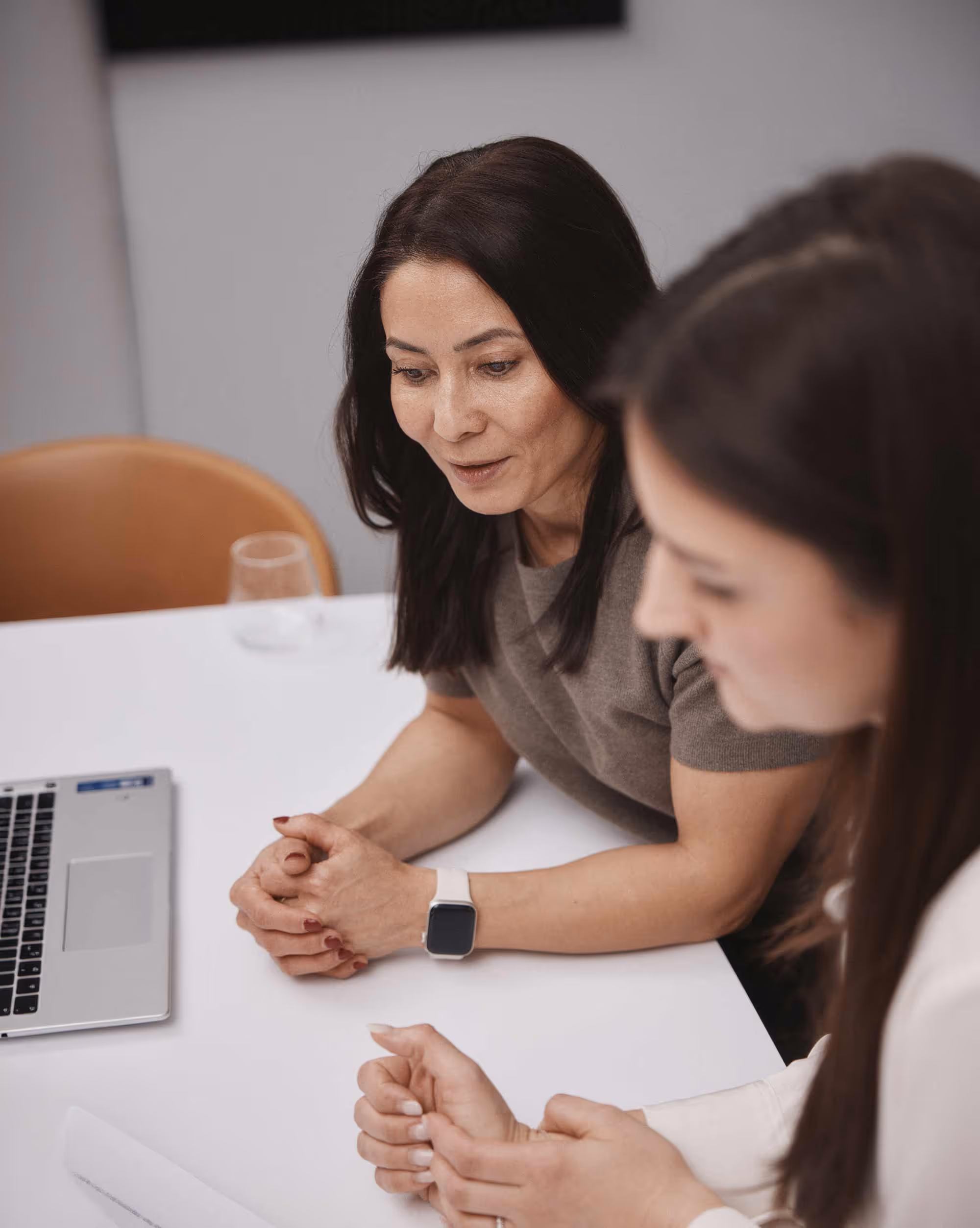 Två kvinnor sitter vid ett bord med en bärbar dator och ett dokument, engagerade i samtal.