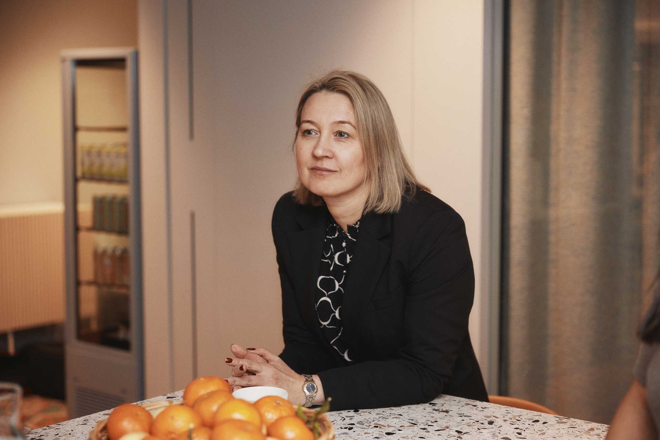 Kvinna med kort blont hår, svart kavaj och mönstrad skjorta sitter vid ett bord med en skål full av apelsiner.