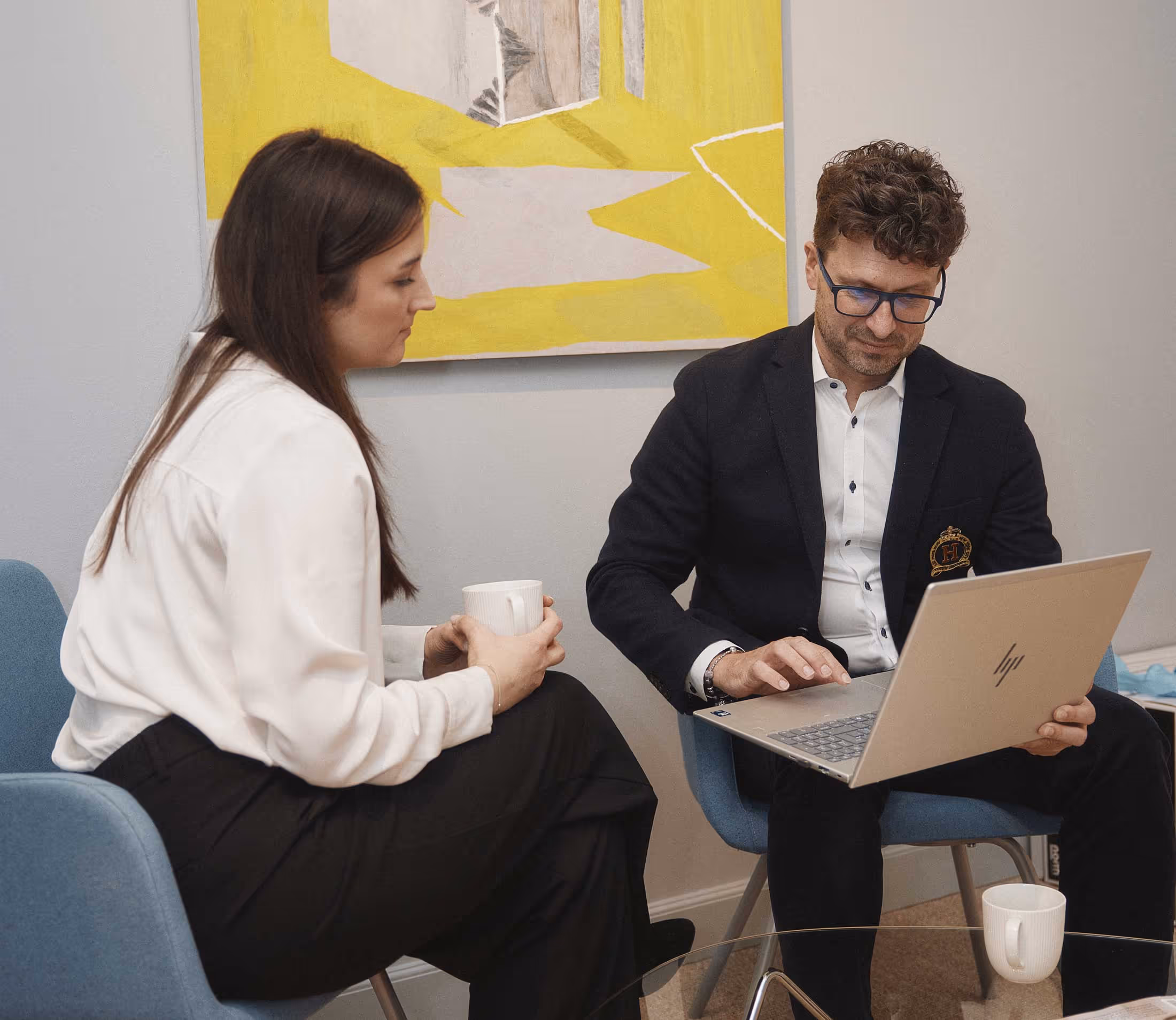 Två personer sitter i blå fåtöljer, en kvinna med vit blus och svart byxor håller en mugg medan en man med glasögon och mörk kavaj arbetar på en bärbar dator.