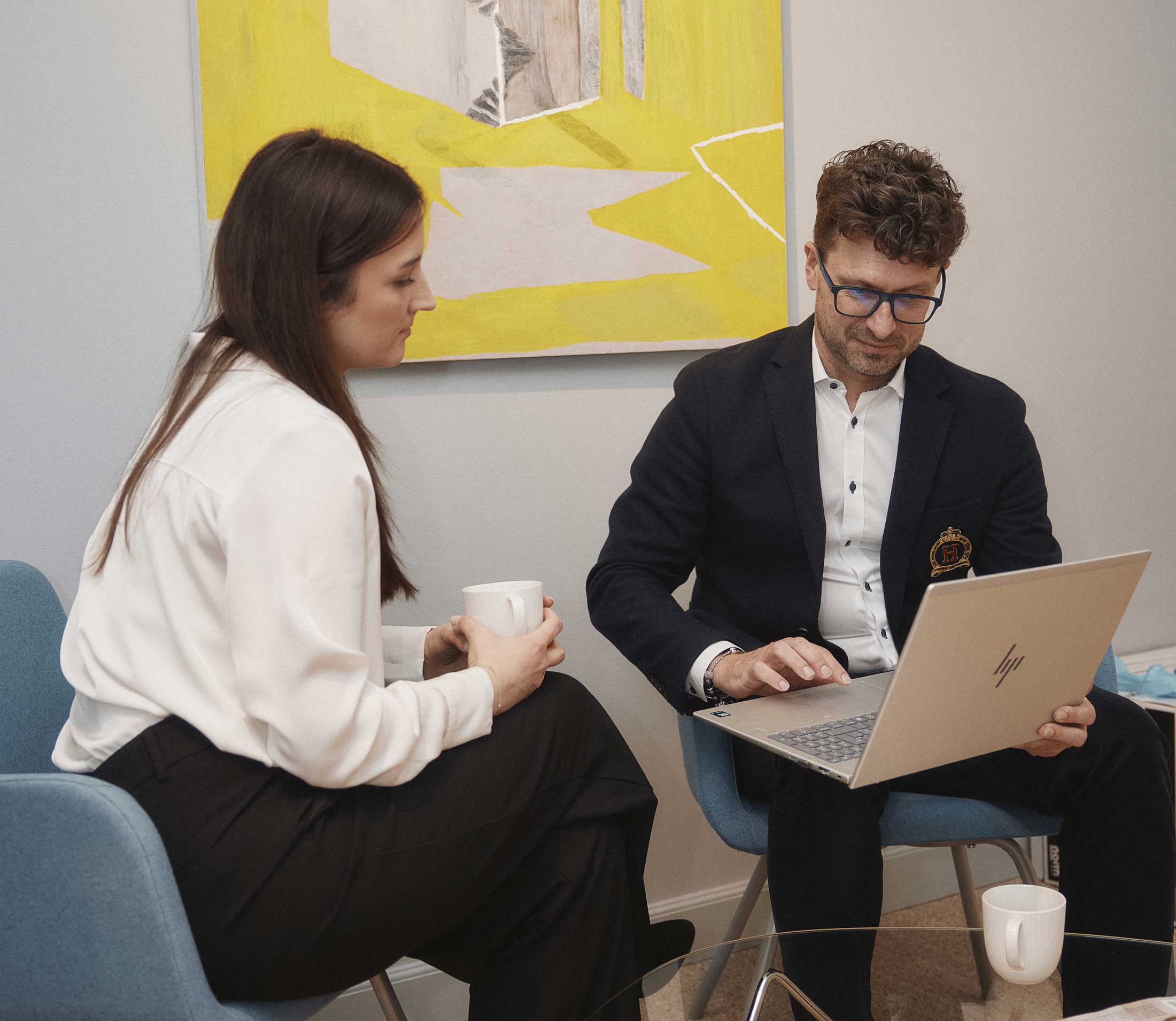 Två personer sitter i blå fåtöljer, en kvinna med vit blus och svart byxor håller en mugg medan en man med glasögon och mörk kavaj arbetar på en bärbar dator.