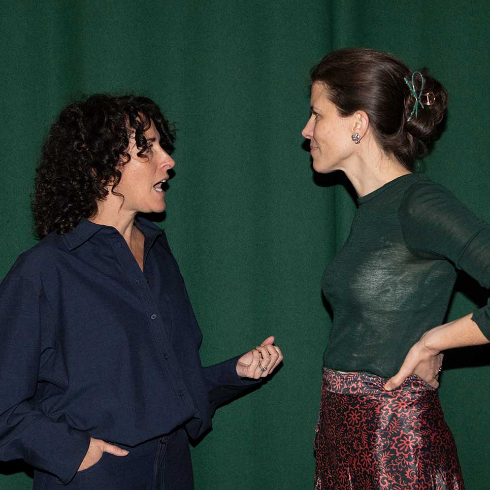 Two women standing and conversing in front of a green backdrop, one with curly hair speaking and the other with a bun listening attentively.