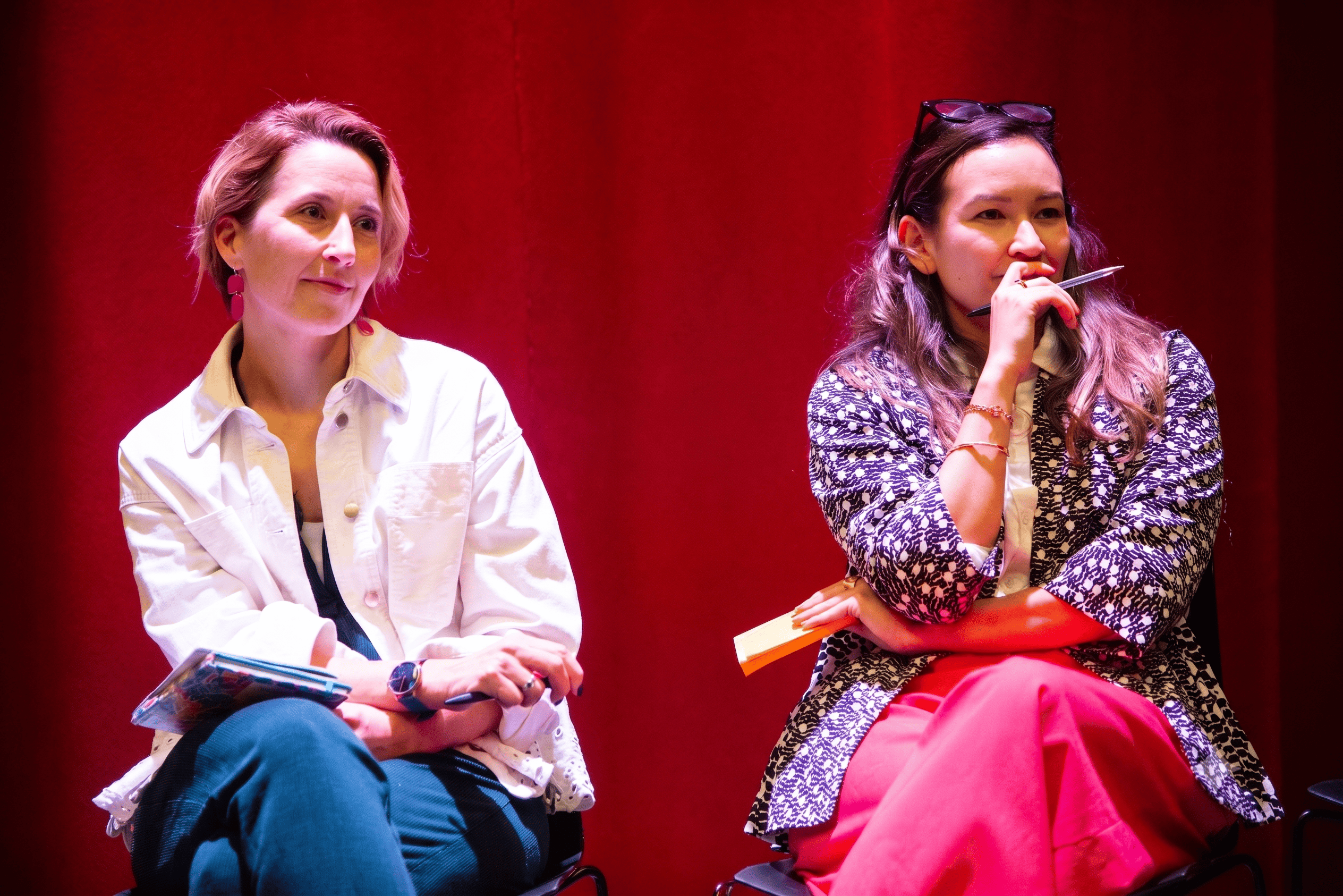 Two women standing and conversing in front of a green backdrop, one with curly hair speaking and the other with a bun listening attentively.