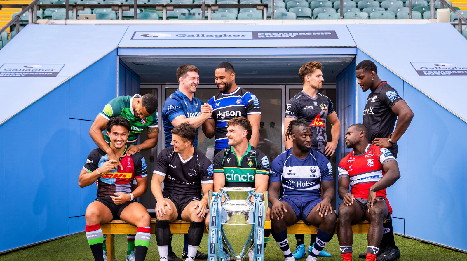 Rugby players in different team jerseys posing with a championship trophy at stadium entrance.
