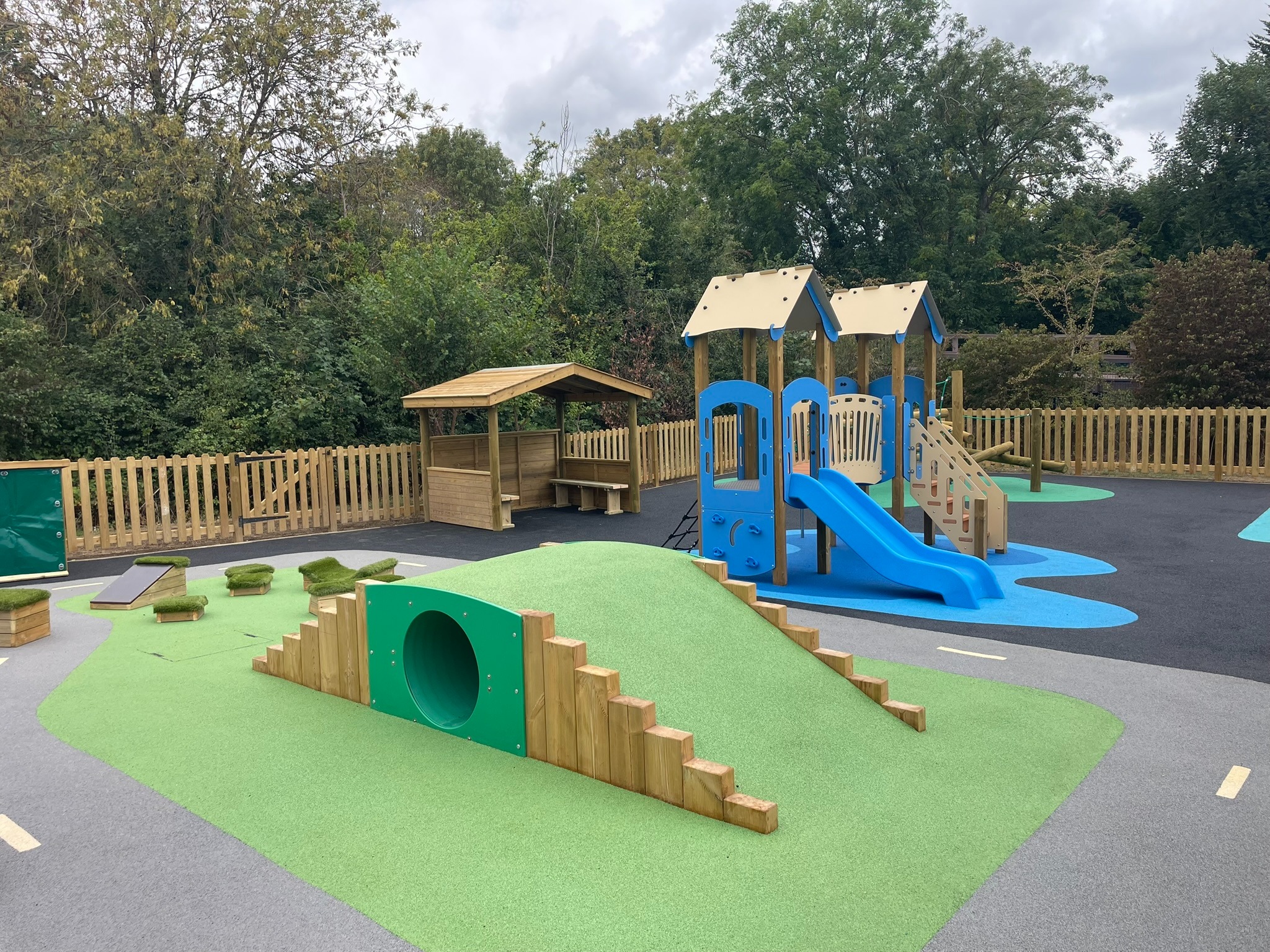 Blue twin-tower playground and slide beside a grass-covered mound with a tunnel, timber shelter and colourful surfacing