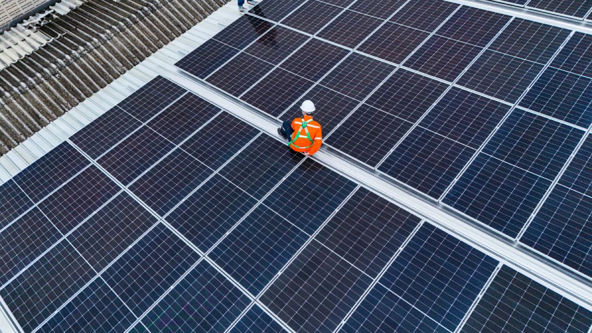 Handwerker sitzt zwischen zwei Reihen an Solarmodulen auf dem Dach einer Fabrik