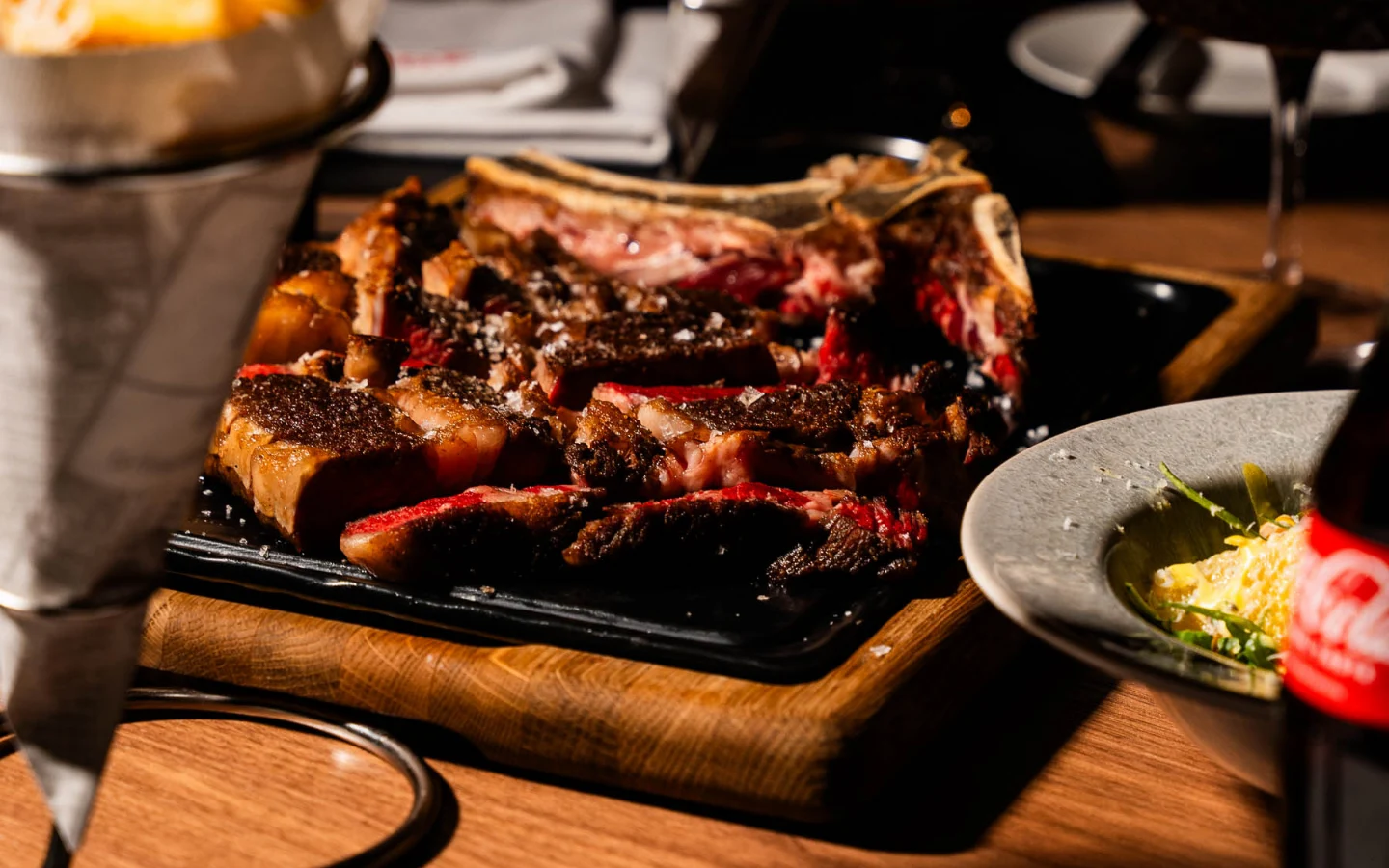 Aufgeschnittenes Tomahawk-Steak auf schwarzer Platte mit Beilagen und Getränken auf gedecktem Tisch.