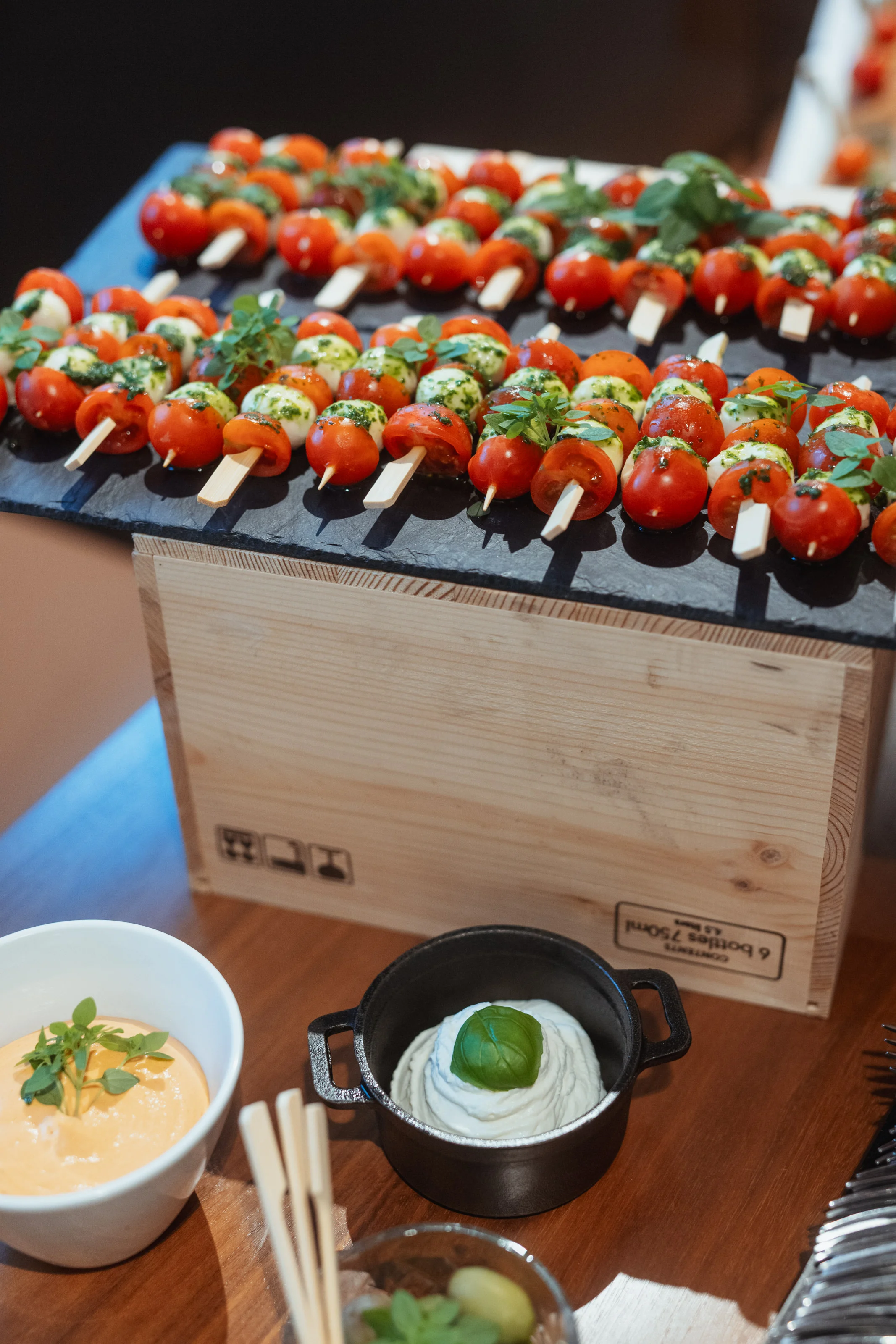 Ein Teller mit zahlreichen Caprese-Spießen, die auf einer dunklen Schieferplatte präsentiert werden. Jeder Spieß ist mit kleinen, runden Tomaten, Mozzarellakugeln und frischen Basilikumblättern bestückt und mit einer grünen Pesto- oder Kräutersoße beträufelt.