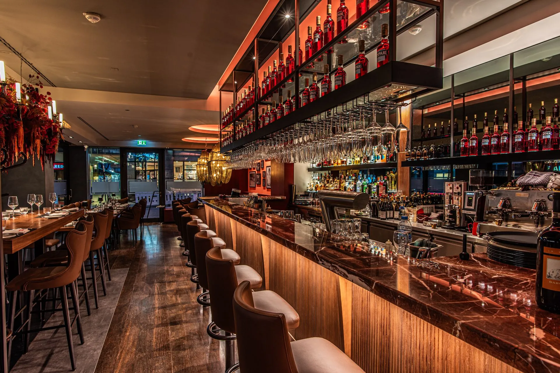 Blick in eine moderne, stilvolle Bar mit einer langen Theke, an der eine Reihe von Barhockern steht. Hinter der Theke sind zahlreiche Spirituosenflaschen auf beleuchteten Regalen angeordnet und Weingläser hängen über Kopf.