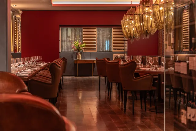 Ein eleganter, langer Esstisch ist in einem Restaurant mit einem warmen, roten Ambiente aufgebaut. Auf beiden Seiten des Tisches stehen braune, lederne Stühle. Im Hintergrund gibt es eine Kommode mit einer Vase voller Blumen und eine Wand mit Holzpaneelen. Der Boden besteht aus poliertem Holzboden, der das Licht reflektiert.