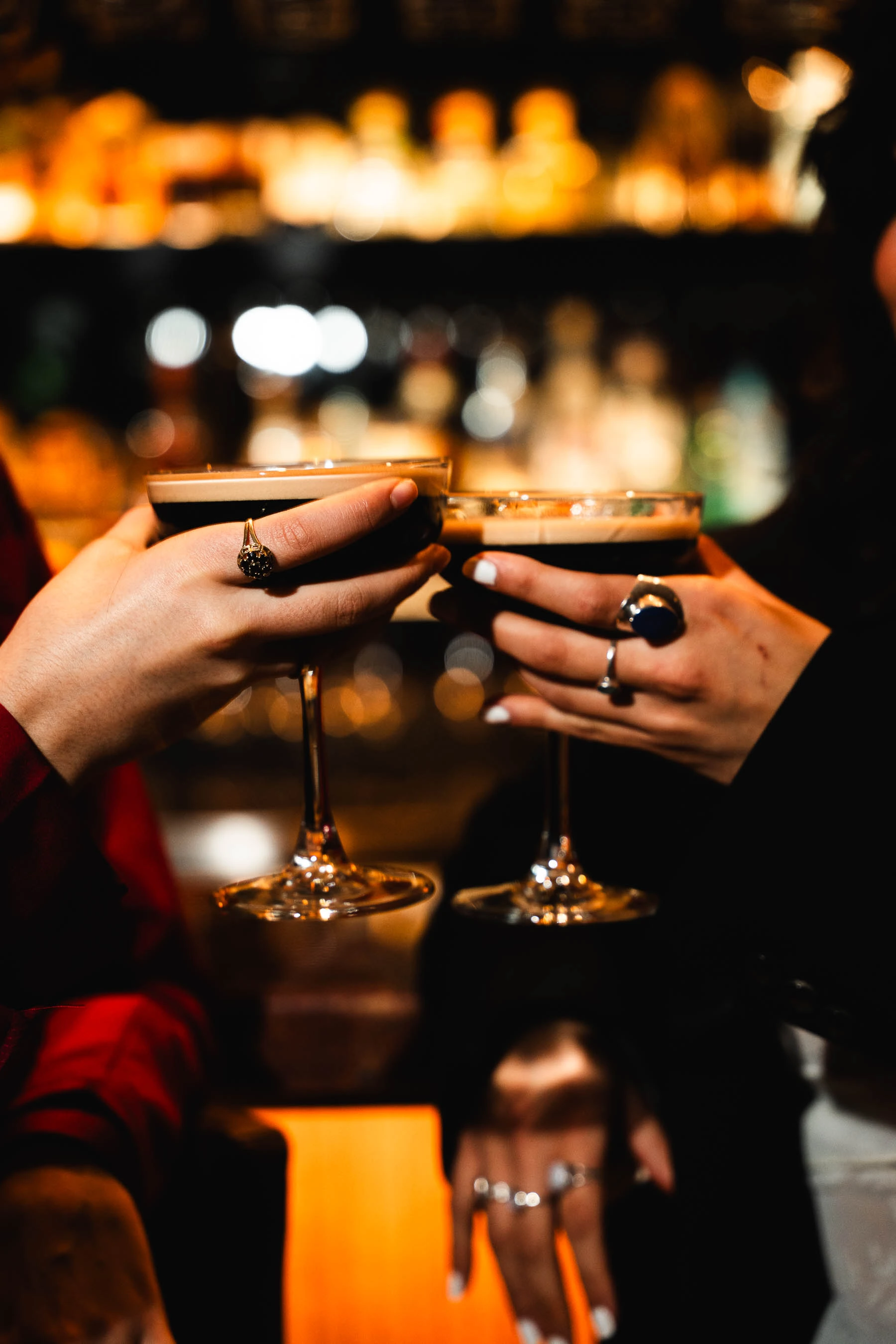 Zwei Personen stoßen mit eleganten Martinigläsern, die dunkle Cocktails mit Schaumkrone enthalten, an. Die Hände der Personen sind mit markanten Ringen geschmückt und der Hintergrund einer Bar ist unscharf.