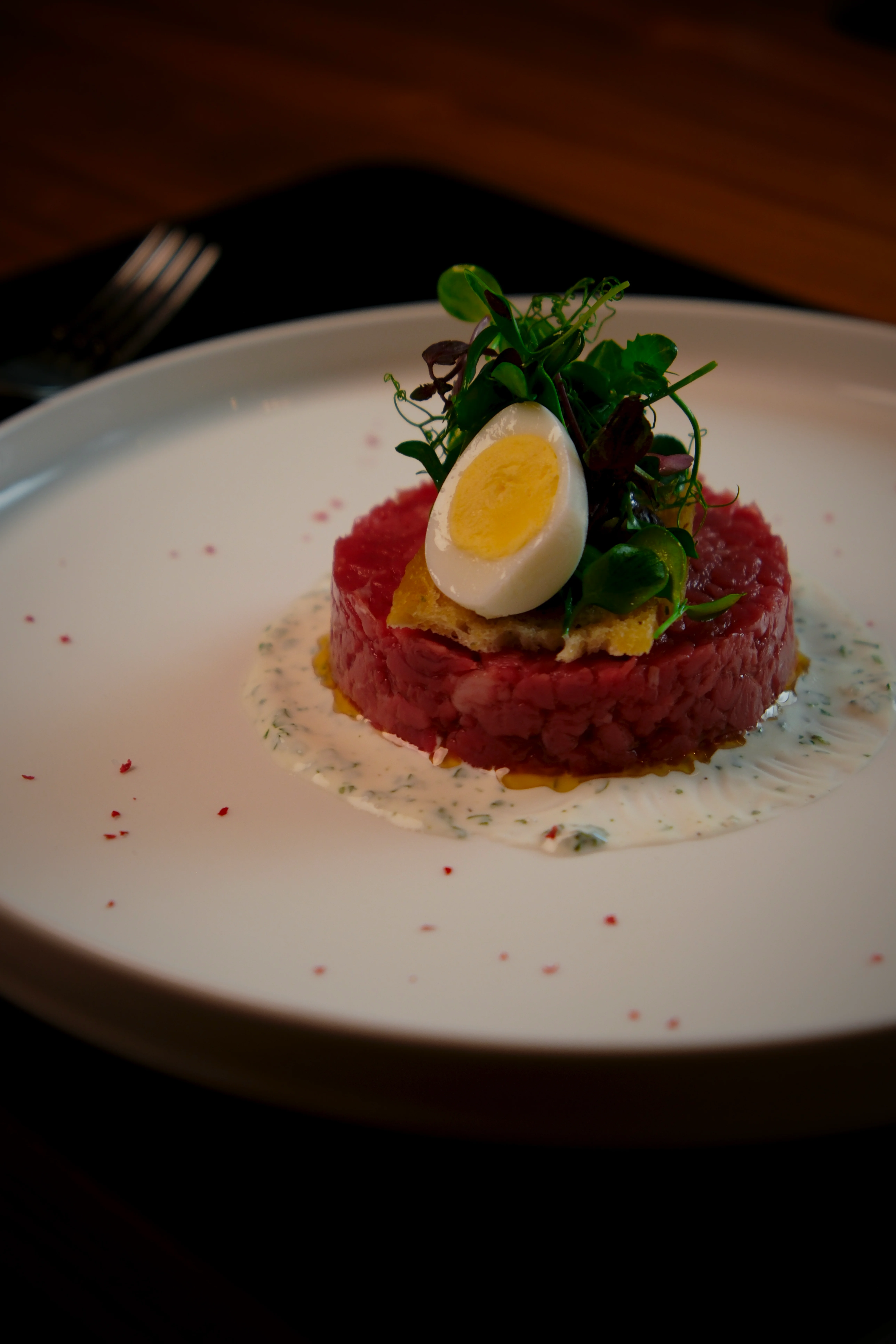 Fein angerichtetes Rindertatar auf einem Spiegel aus Kräuter-Sauce, garniert mit einem halben Ei und frischen Kräutern, serviert auf einem weißen Teller.