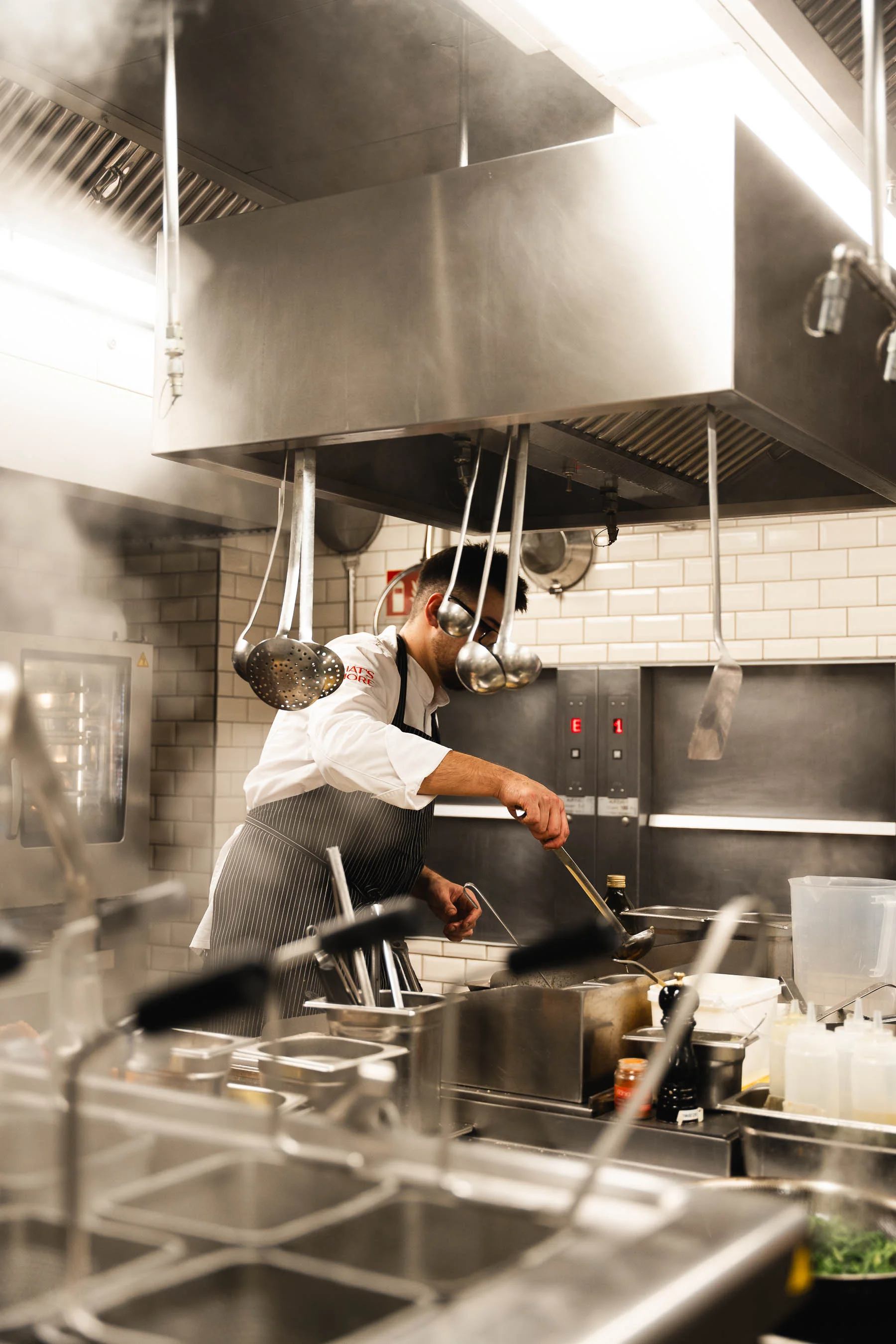 Koch im weißen Outfit mit schwarzer Schürze arbeitet am Herd in einer professionellen Restaurantküche, während Dampf aufsteigt.