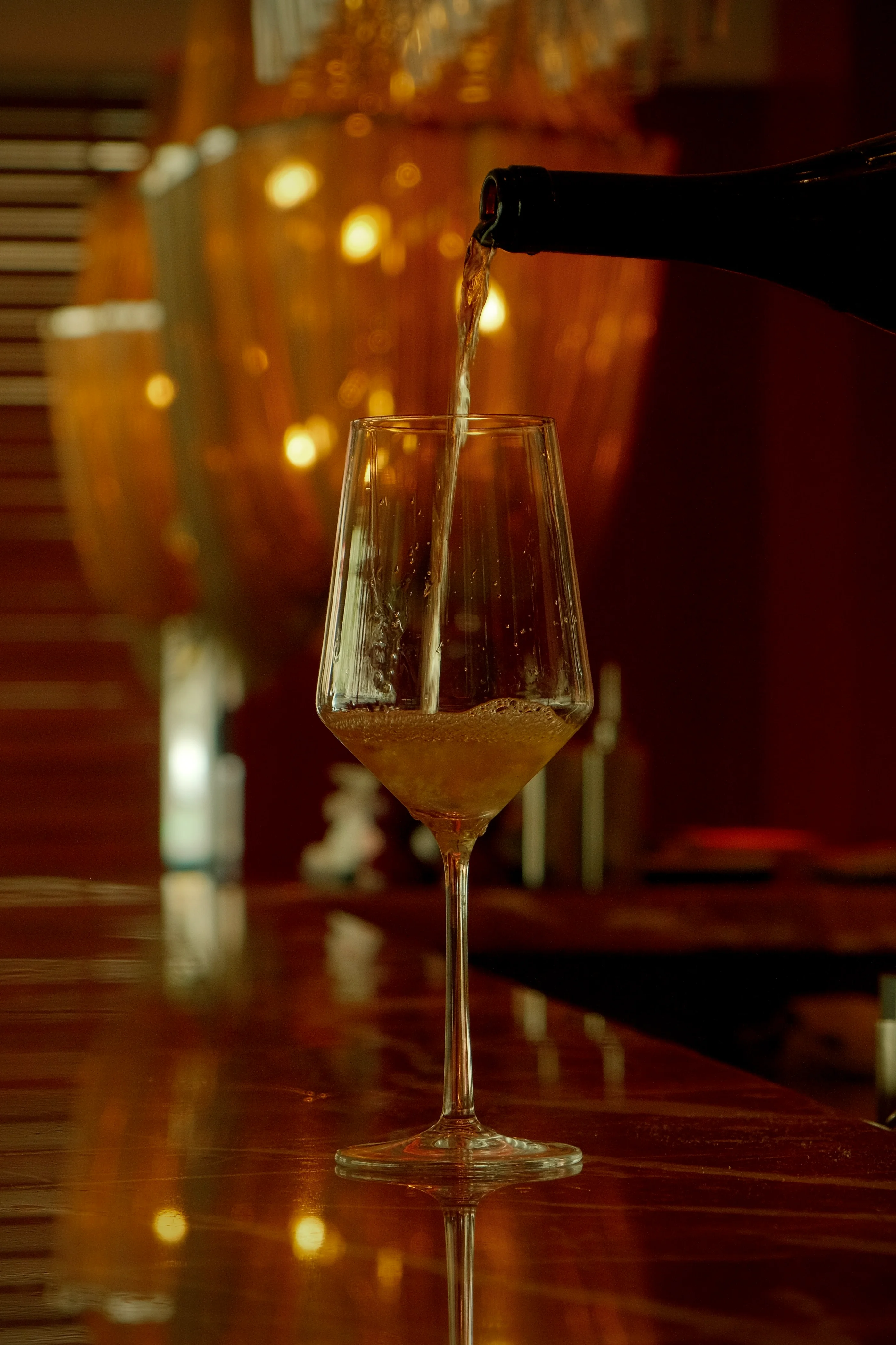 Nahaufnahme eines Weinglases, in das Weißwein an einer stilvollen Bar mit goldenen Lichtern eingeschenkt wird.