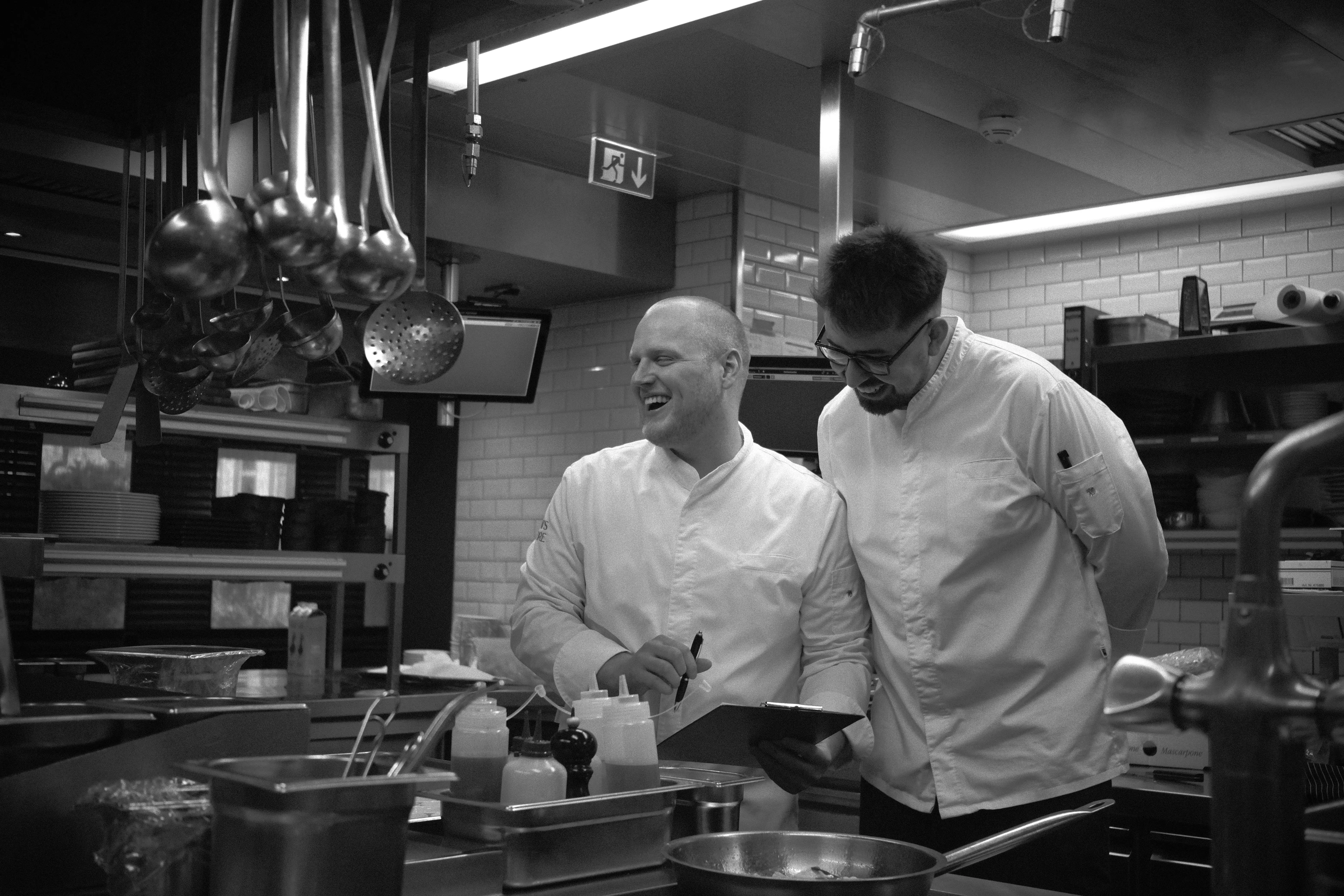 Zwei Köche in weißem Outfit lachen und besprechen sich in der professionellen Küche, umgeben von Kochutensilien und Hängeschöpflöffeln.