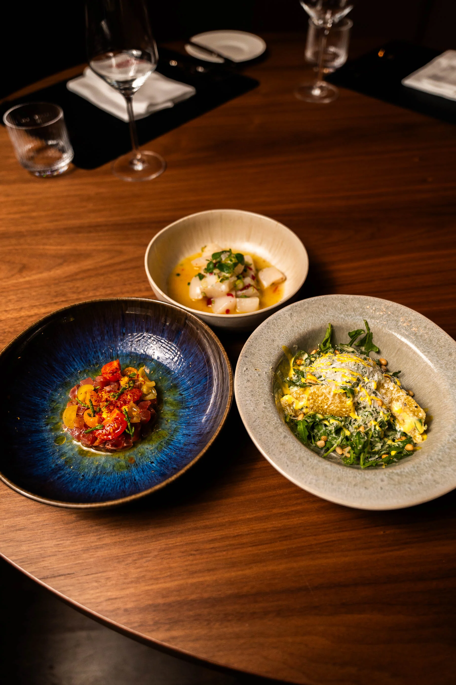 Drei stilvoll angerichtete Vorspeisen in Keramikschalen auf einem Holztisch, umgeben von Gläsern und elegant gedecktem Restaurantambiente.