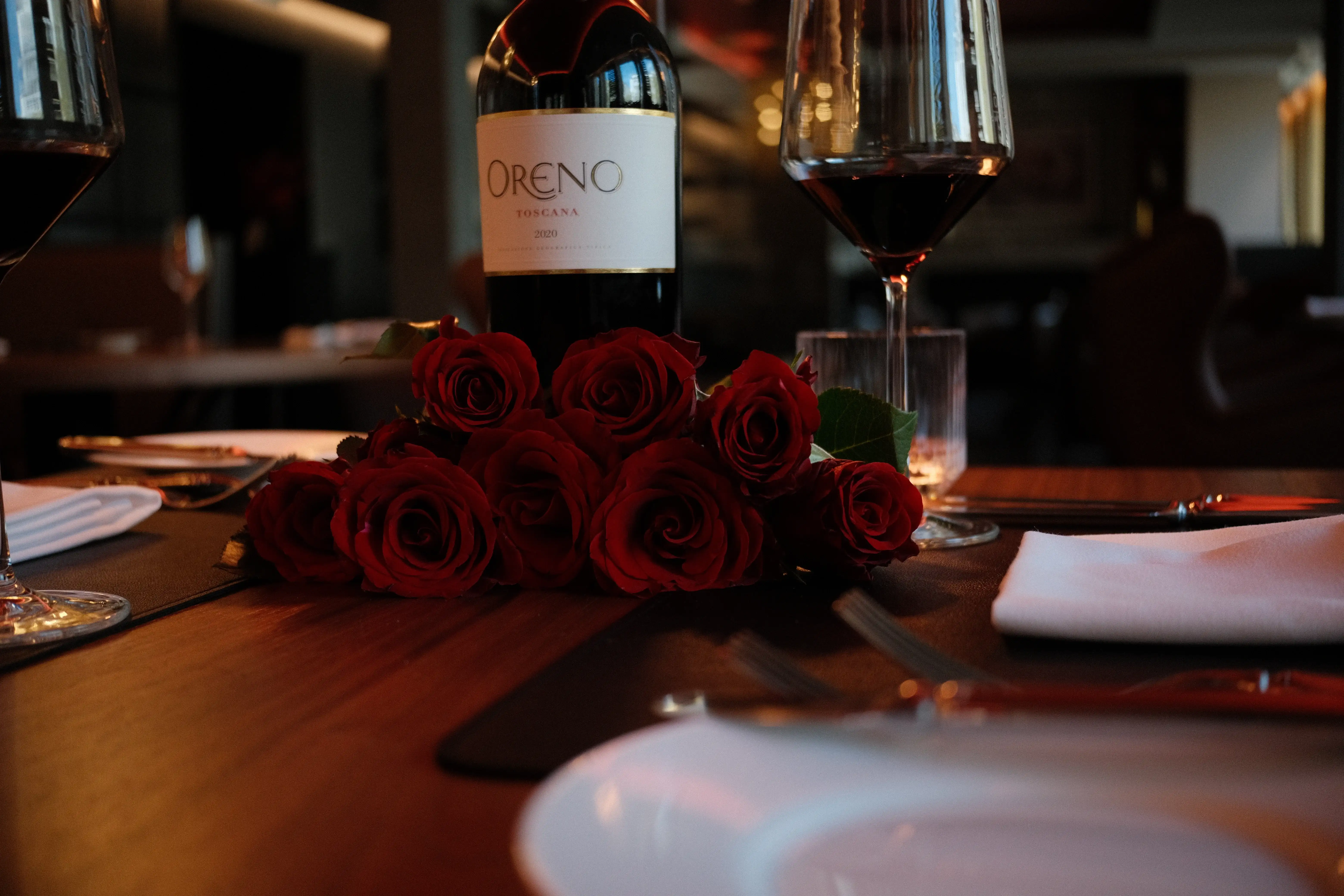 Nahaufnahme einer Rotweinflasche mit zwei Gläsern und einem Strauß roter Rosen als romantische Tischdekoration.