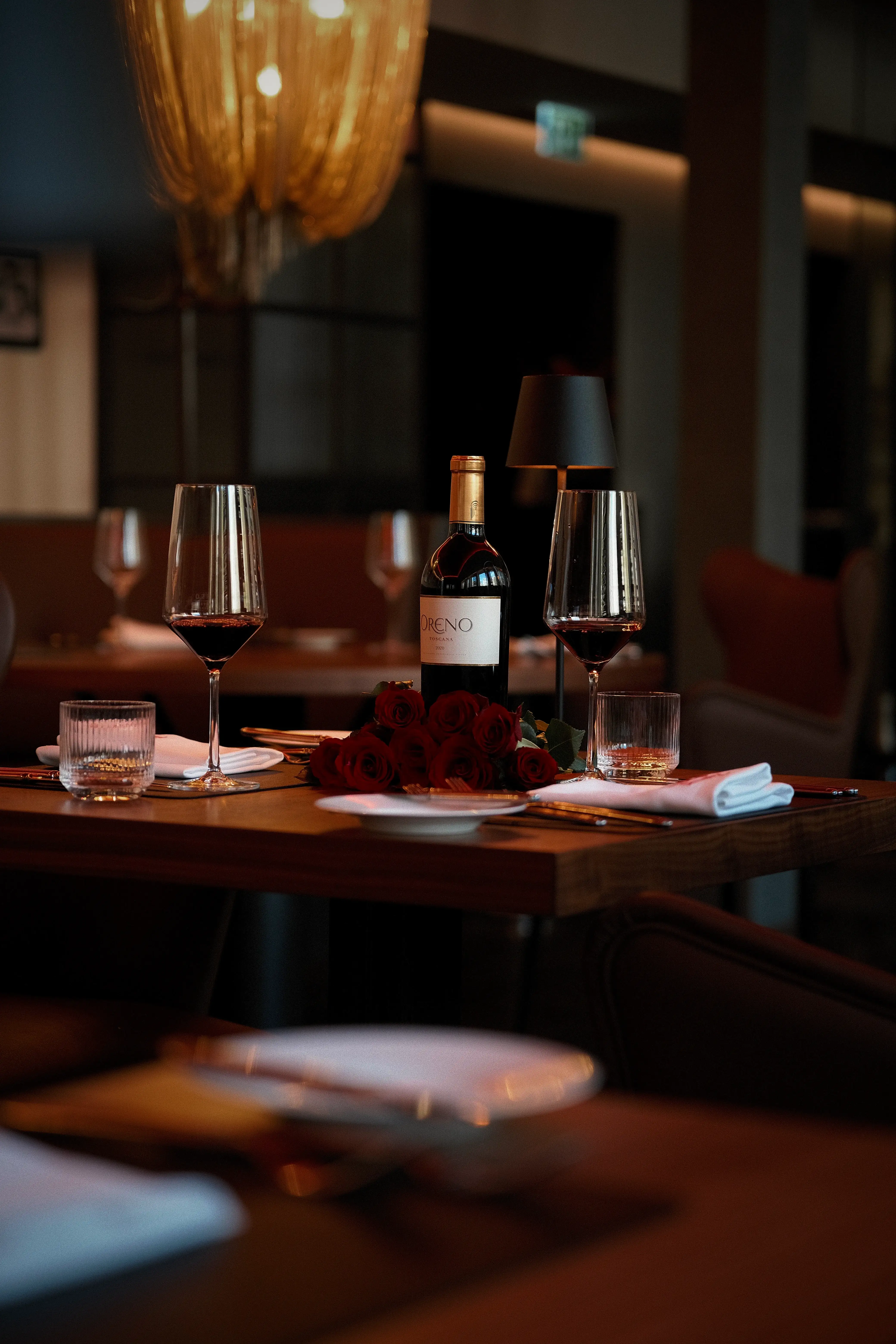 Festlich gedeckter Tisch mit einer Flasche Rotwein, zwei Gläsern und einem Strauß roter Rosen, stimmungsvoll beleuchtet im Restaurant.