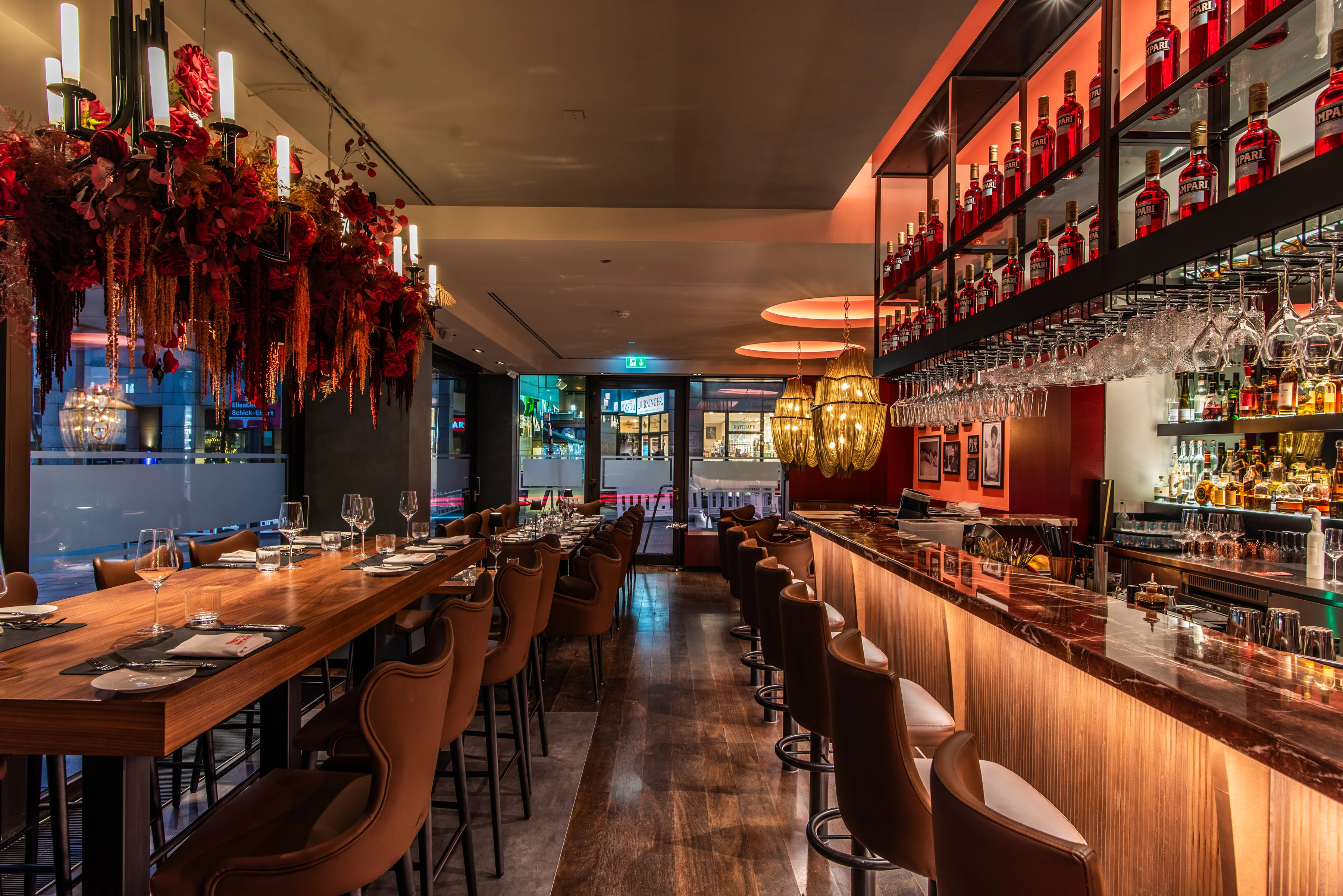 Bar-Szene bei Nacht. Der Hauptfokus liegt auf einer langen, beleuchteten Bar mit einer Theke aus poliertem Marmor, an der mehrere hohe, braune Ledersessel stehen. Hinter der Bar befindet sich ein Regal mit zahlreichen Flaschen und Gläsern, und von der Decke hängen moderne Kronleuchter herab. Auf der linken Seite erstreckt sich ein langer Holztisch, über dem eine aufwendige Dekoration aus roten, getrockneten Blumen und Kerzen hängt.