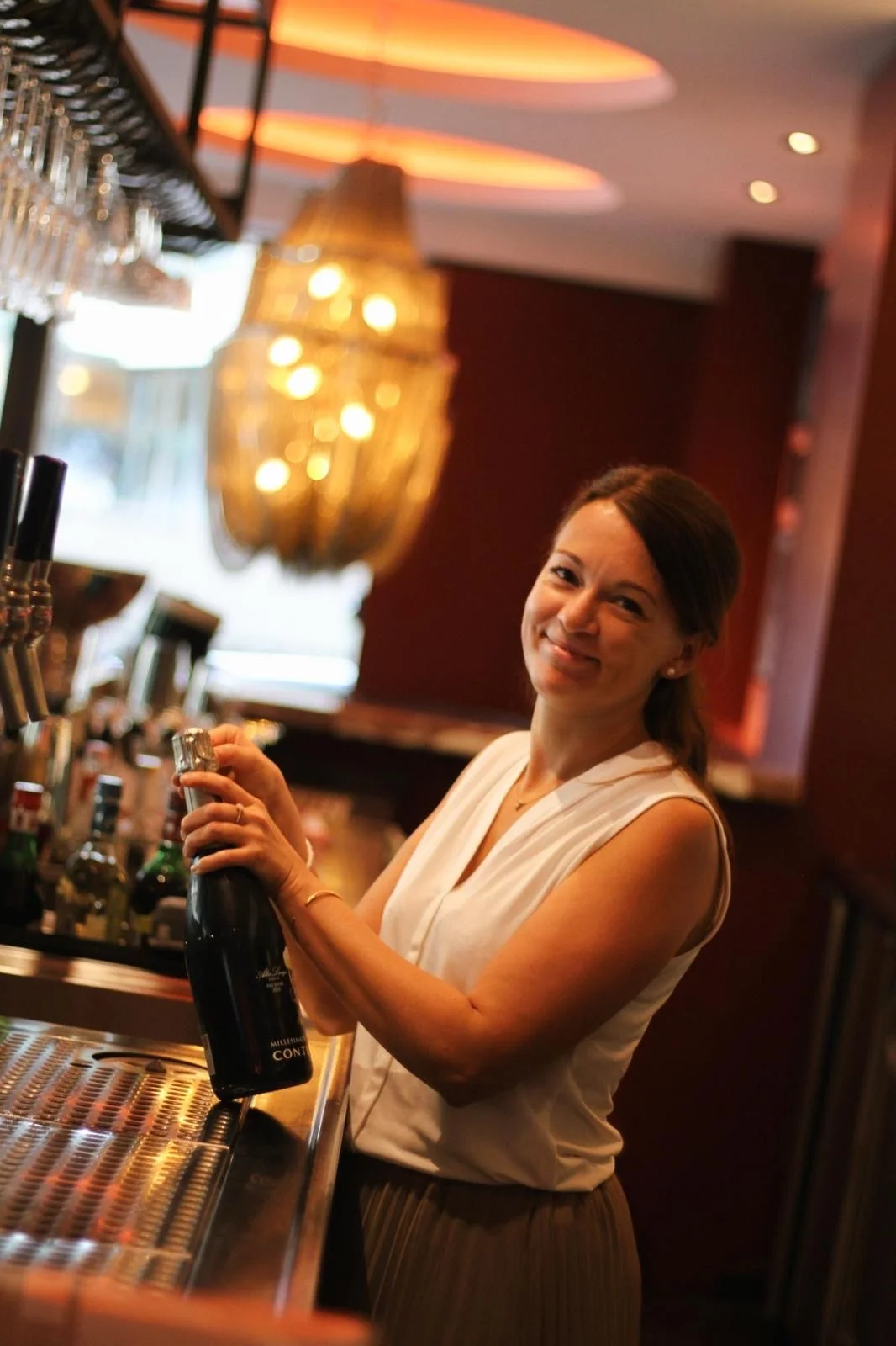 Freundliche Barkeeperin im That’s Amore Stuttgart öffnet eine Flasche Sekt an der stilvoll beleuchteten Bar mit goldenen Lichtern im Hintergrund.
