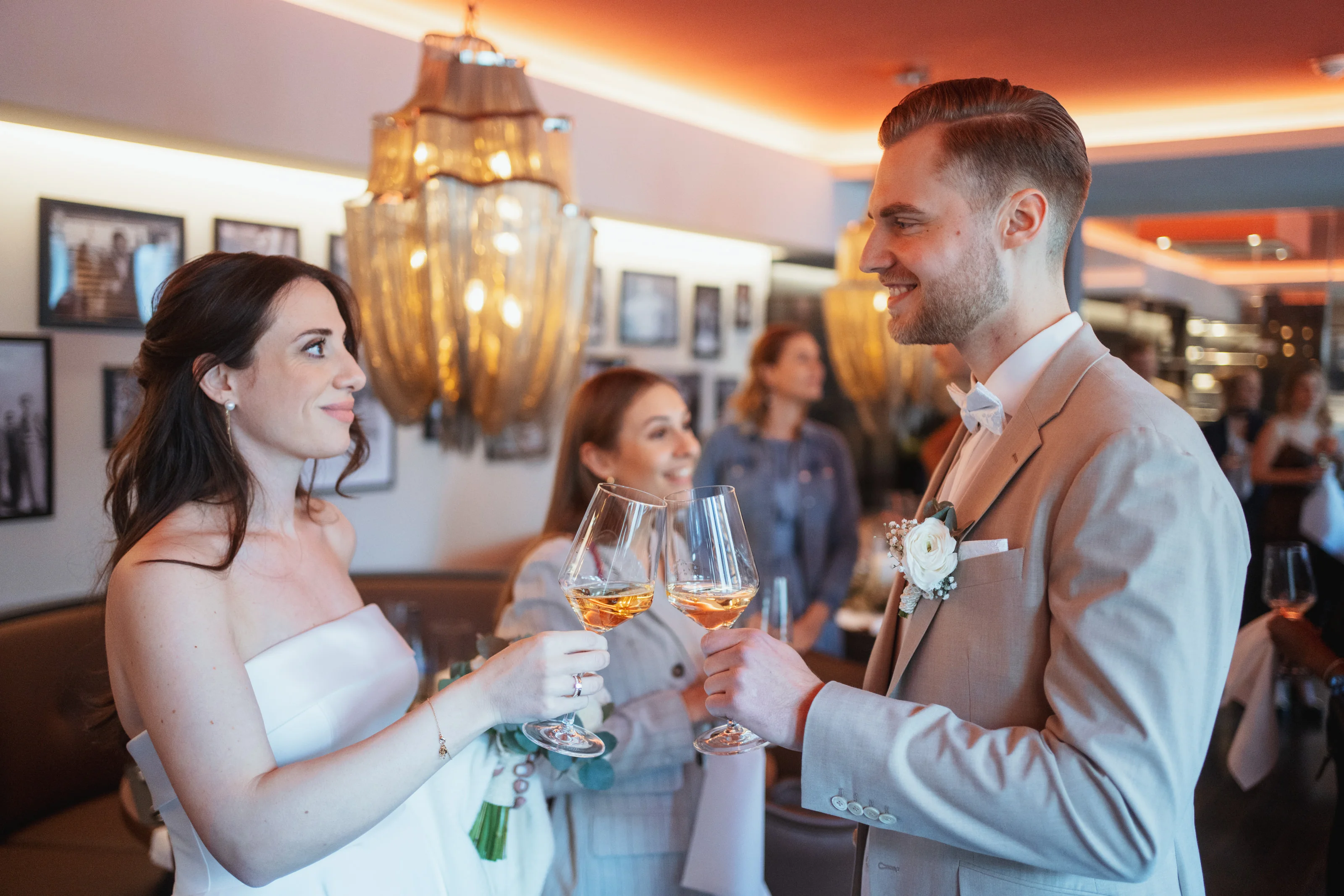 Braut und Bräutigam stoßen mit Weingläsern an und lächeln sich an, im Hintergrund sind Gäste und eine elegante Lampe zu sehen.