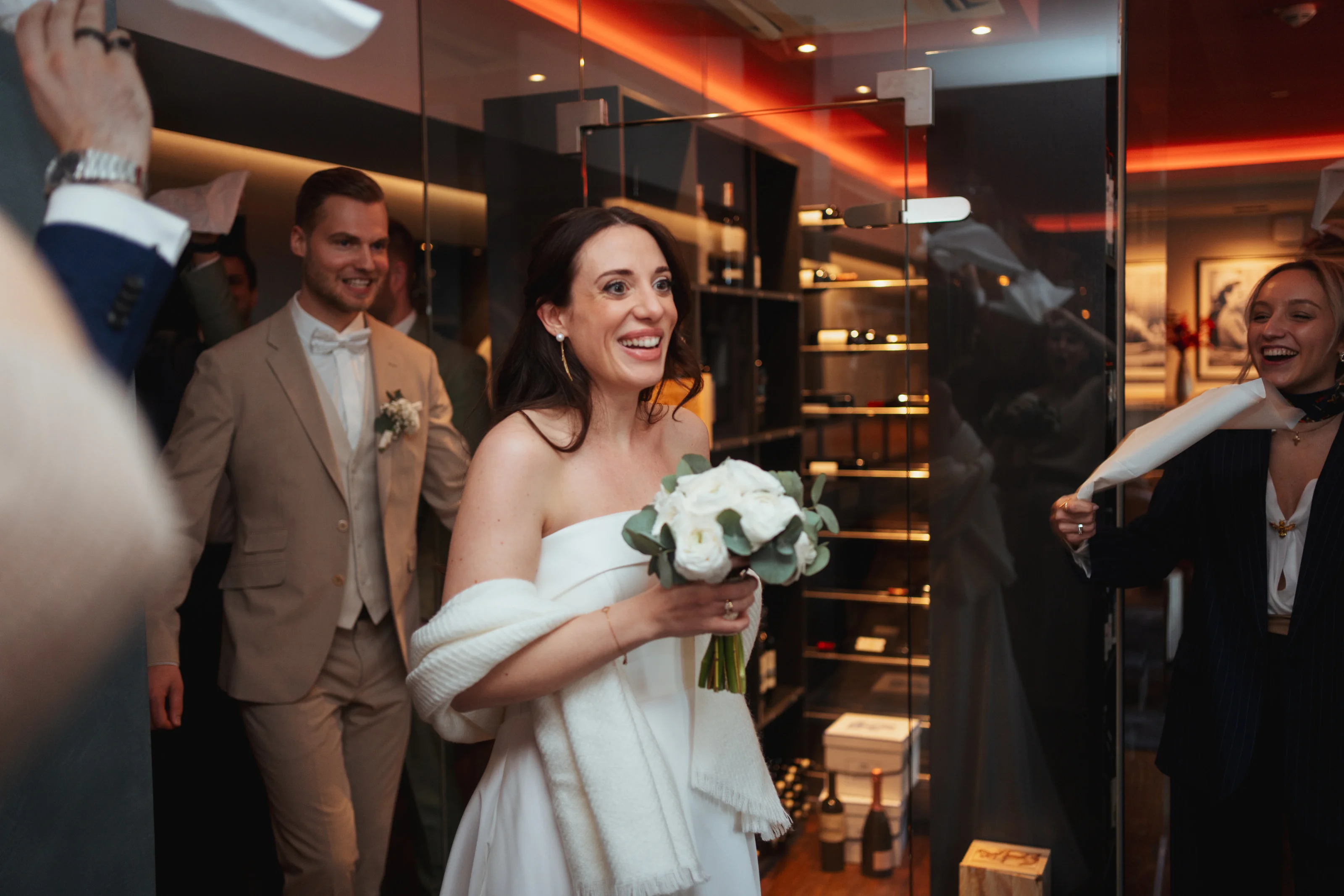 Lächelnde Braut im weißen Kleid mit Blumenstrauß in der Hand betritt den Raum, begleitet vom Bräutigam und jubelnden Gästen.