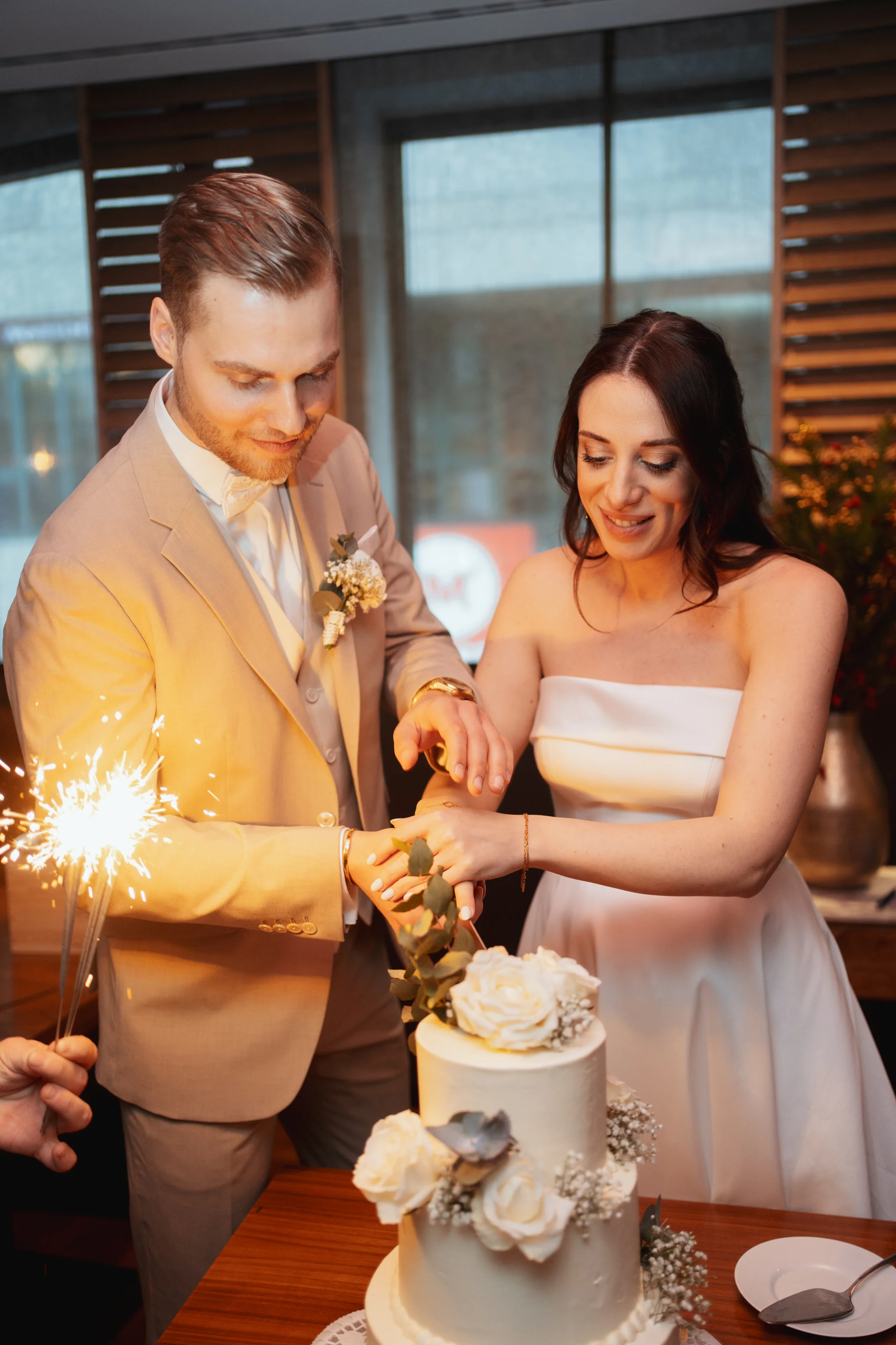 Braut und Bräutigam schneiden gemeinsam eine weiße Hochzeitstorte mit Rosen und Eukalyptus.