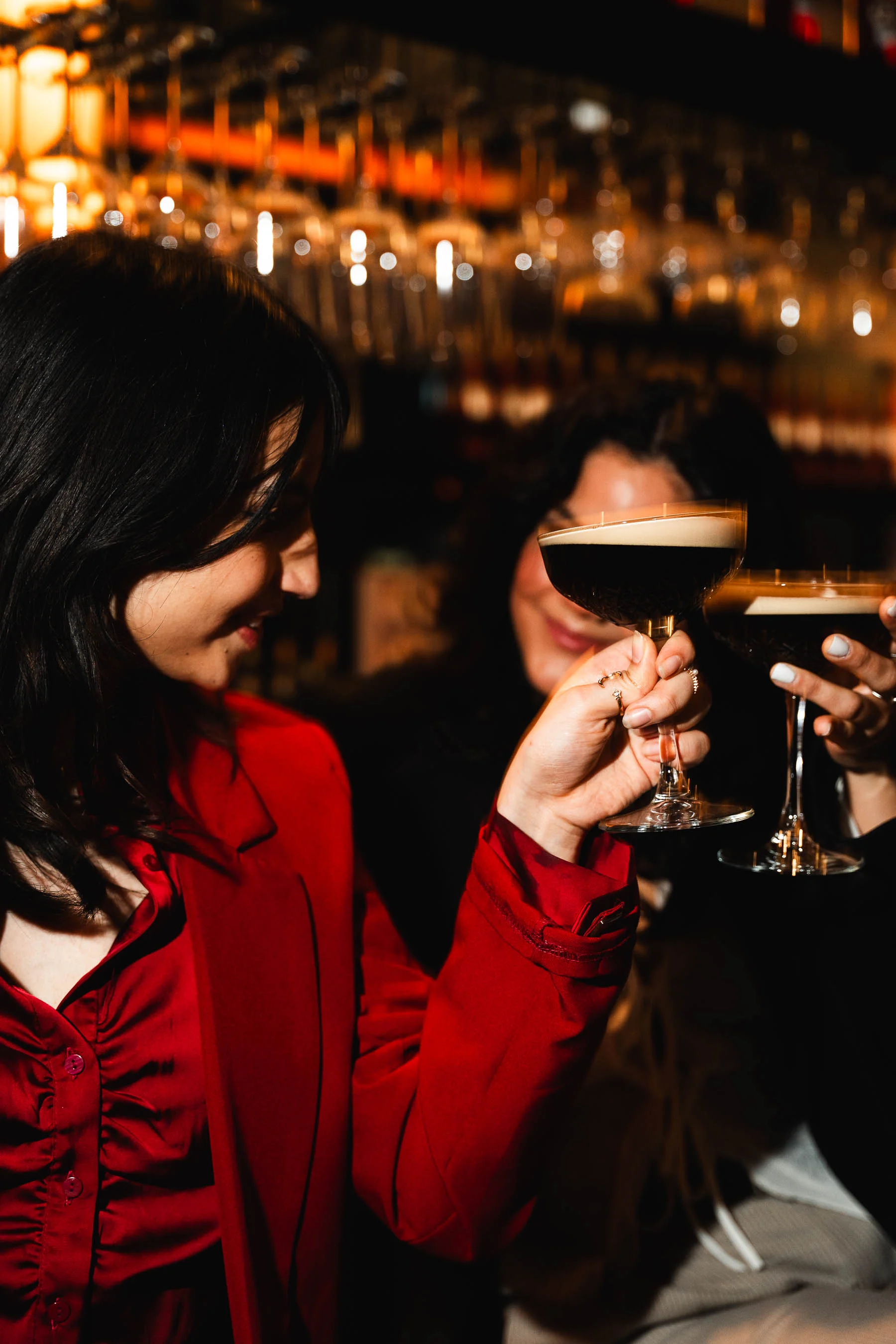 Frau im roten Outfit stößt in einer Bar mit einem Cocktailglas an, im Hintergrund weitere Gäste und Gläser.