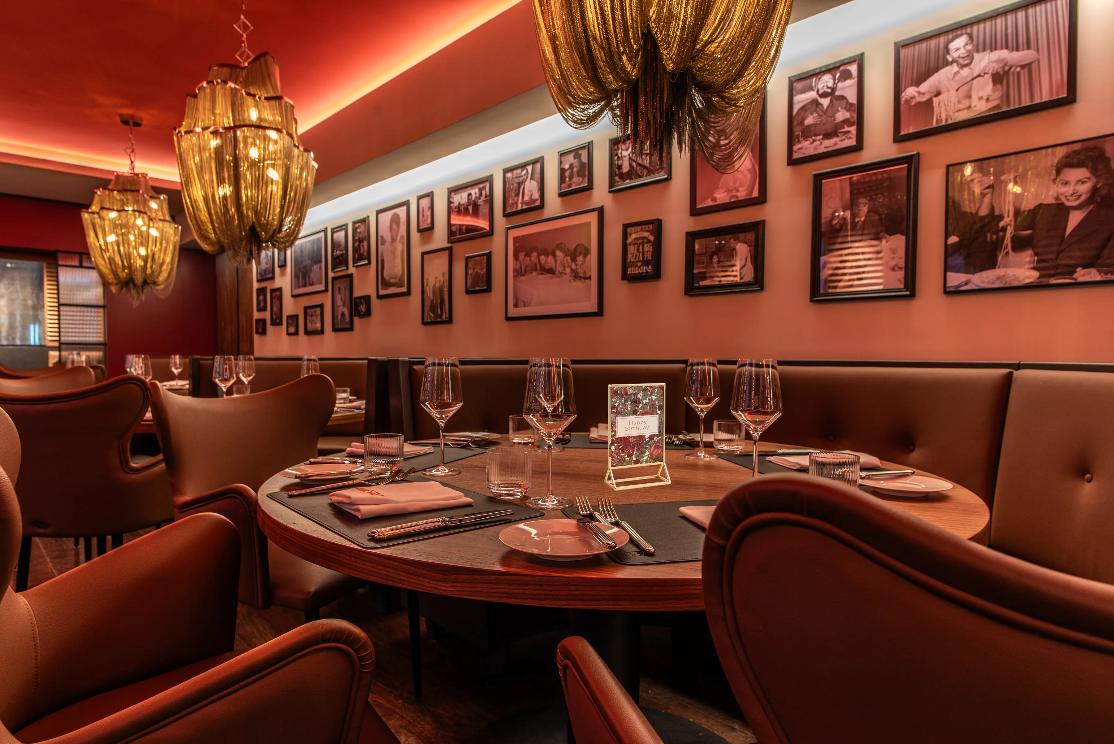 Ein luxuriöses Restaurantinterieur, dominiert von einem großen, runden Tisch, der für mehrere Personen eingedeckt ist. Der Raum ist in warmen Brauntönen und gedämpftem Rot gehalten. An den Wänden hängen zahlreiche gerahmte Fotos in verschiedenen Größen, und von der Decke hängen einzigartige Kronleuchter mit goldenen, kettenartigen Elementen herab.