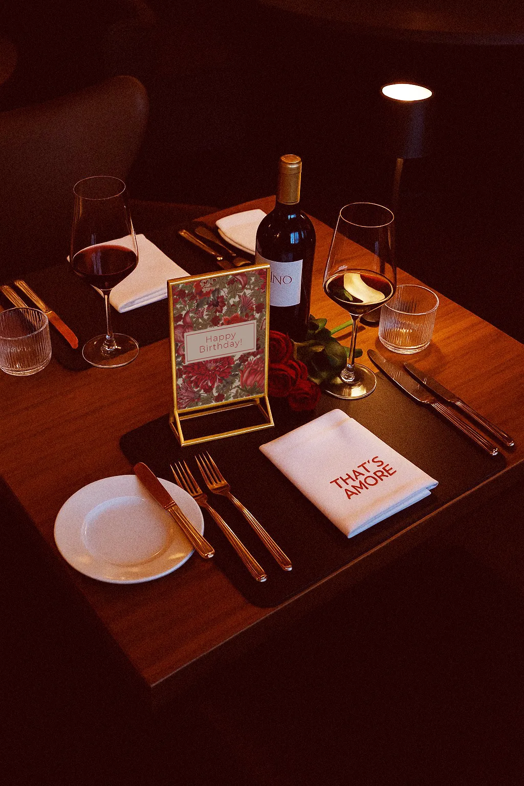Ein intimer, für zwei Personen gedeckter Tisch in einem dunklen Restaurant. Auf dem Holztisch stehen zwei Gläser Rotwein, eine Weinflasche, eine einzelne rote Rose und ein aufgestelltes Schild, das mit der Aufschrift „Happy Birthday!“ und roten Blumenmustern verziert ist. Eine weiße Serviette liegt vor einem Gedeck und ist mit den Worten „THAT'S AMORE!“ bestickt. Die gedämpfte Beleuchtung und die romantischen Elemente schaffen eine warme Atmosphäre.