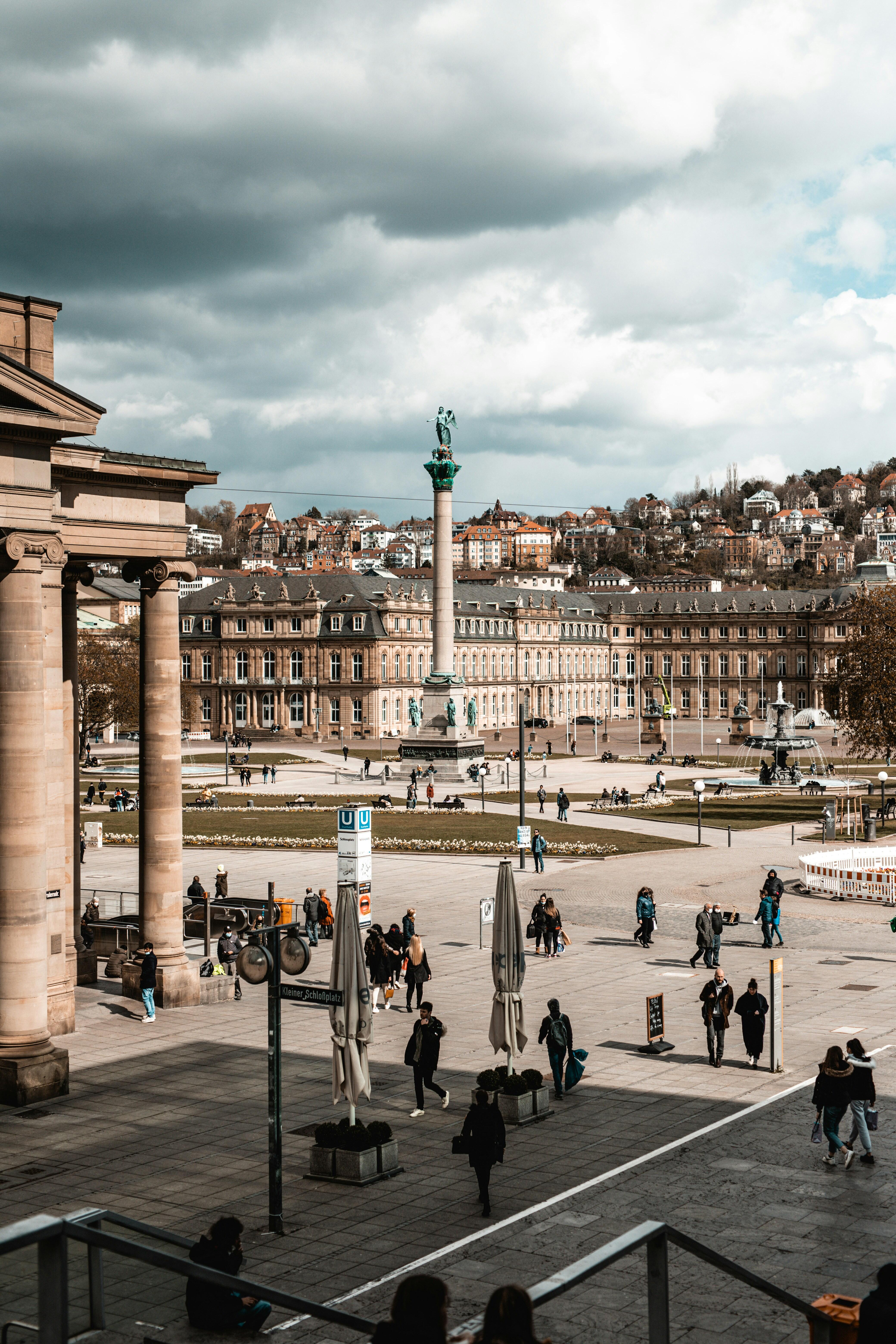 Blick auf den Stuttgarter Schlossplatz mit Jubiläumssäule, historischen Gebäuden und vorbeigehenden Passanten unter dramatischem Himmel.