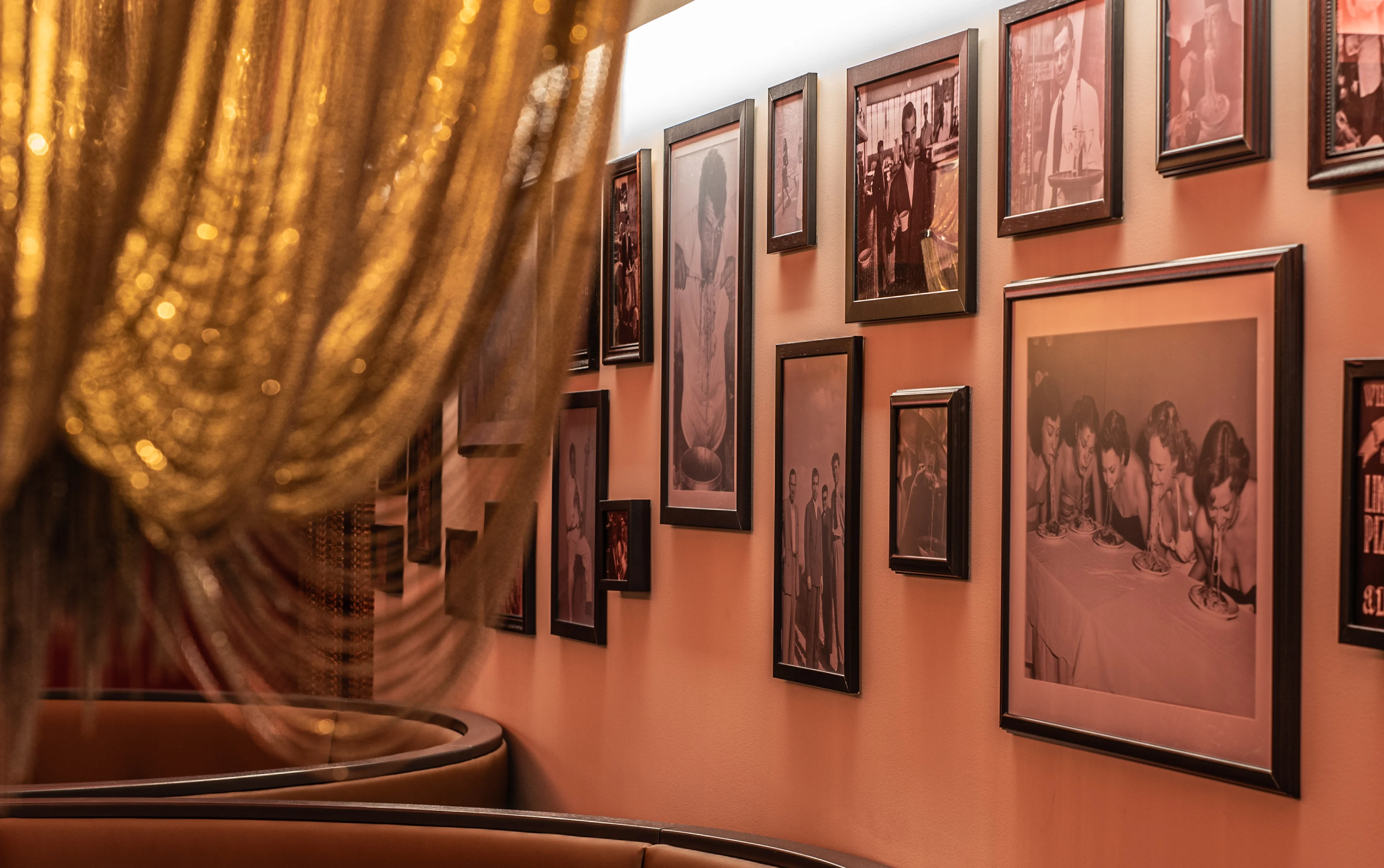 Wand mit mehreren gerahmten Schwarz-Weiß-Fotografien in einem Restaurant, davor runde Sitznischen und eine goldfarbene Lampe im Vordergrund.