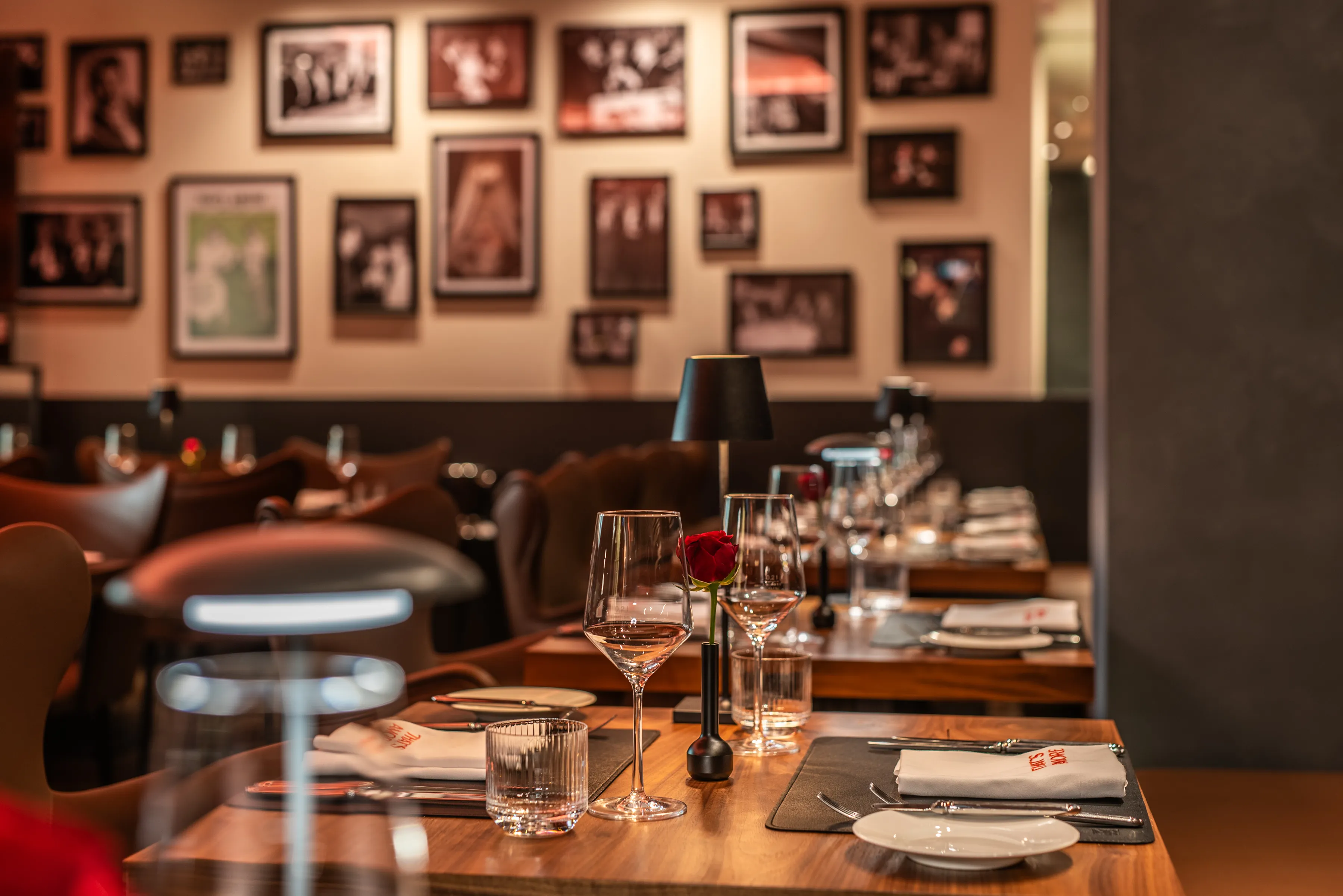 Reihe gedeckter Tische in einem Restaurant mit Weingläsern, Tellern, Servietten und kleinen Tischlampen, im Hintergrund eine Wand mit gerahmten Fotografien.