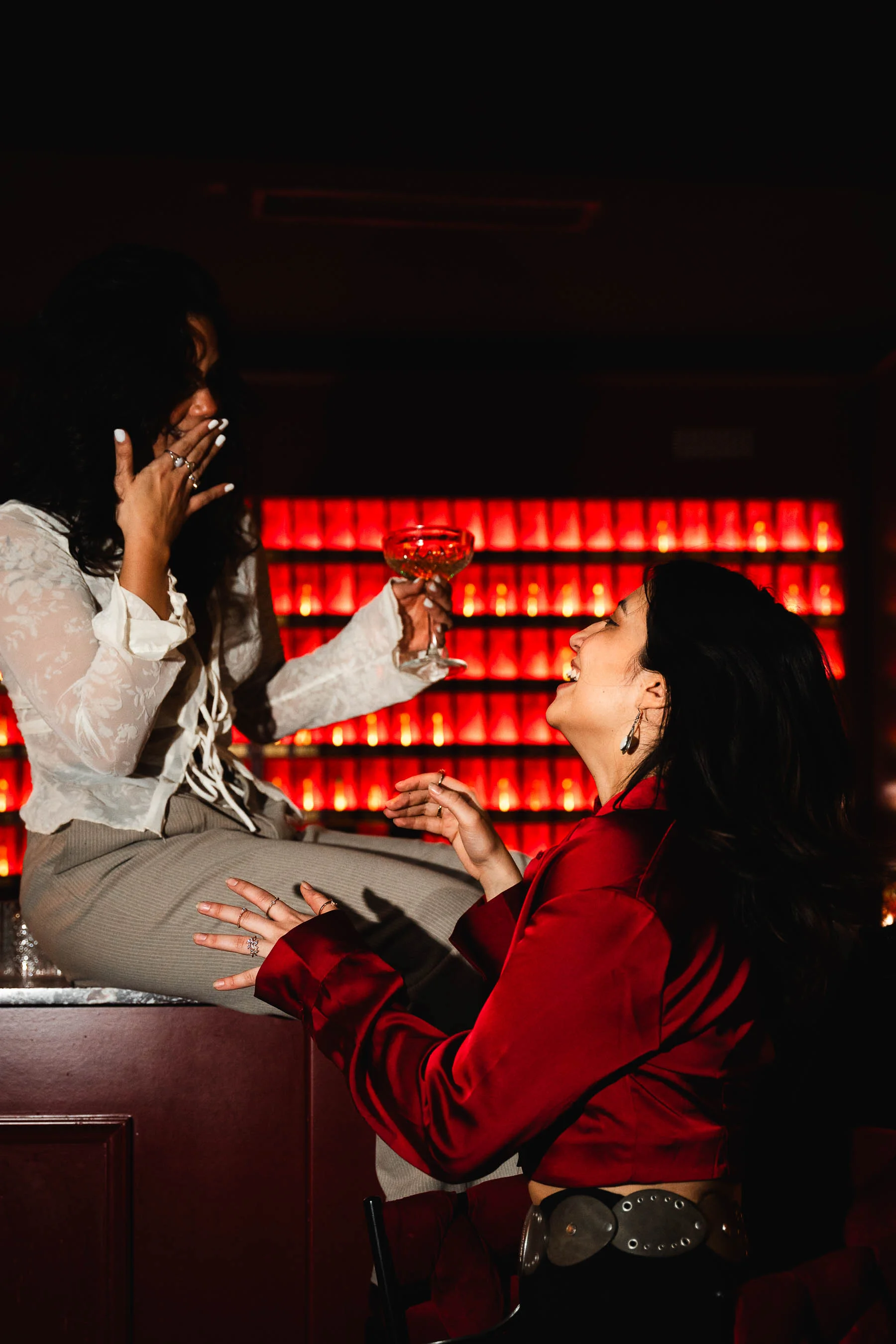 Zwei Frauen lachen gemeinsam in einer Bar mit roter Beleuchtung. Eine sitzt auf der Theke mit einem Cocktailglas in der Hand, die andere steht vor ihr und trägt eine rote Bluse.