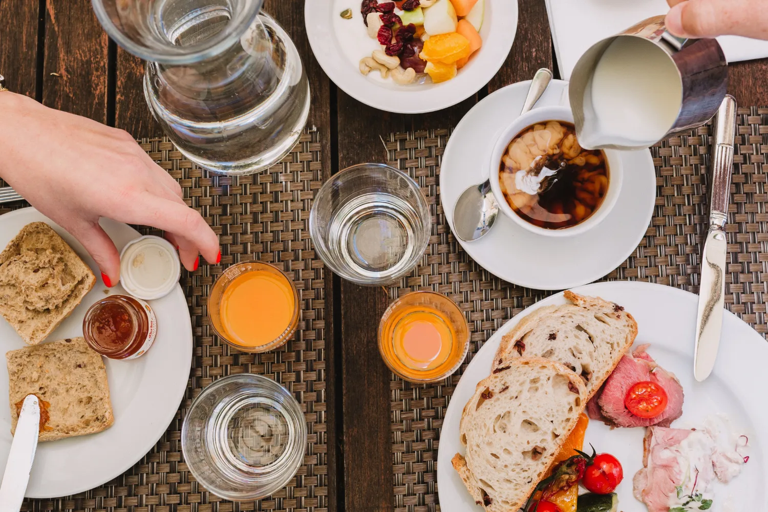 Ein liebevoll gedeckter Frühstückstisch mit frischem Brot, Brötchen, Marmelade, Obstsalat, Aufschnitt, Kaffee mit Milch und Gläsern voller Saft und Wasser. Zwei Personen greifen gerade nach Speisen und Getränken.