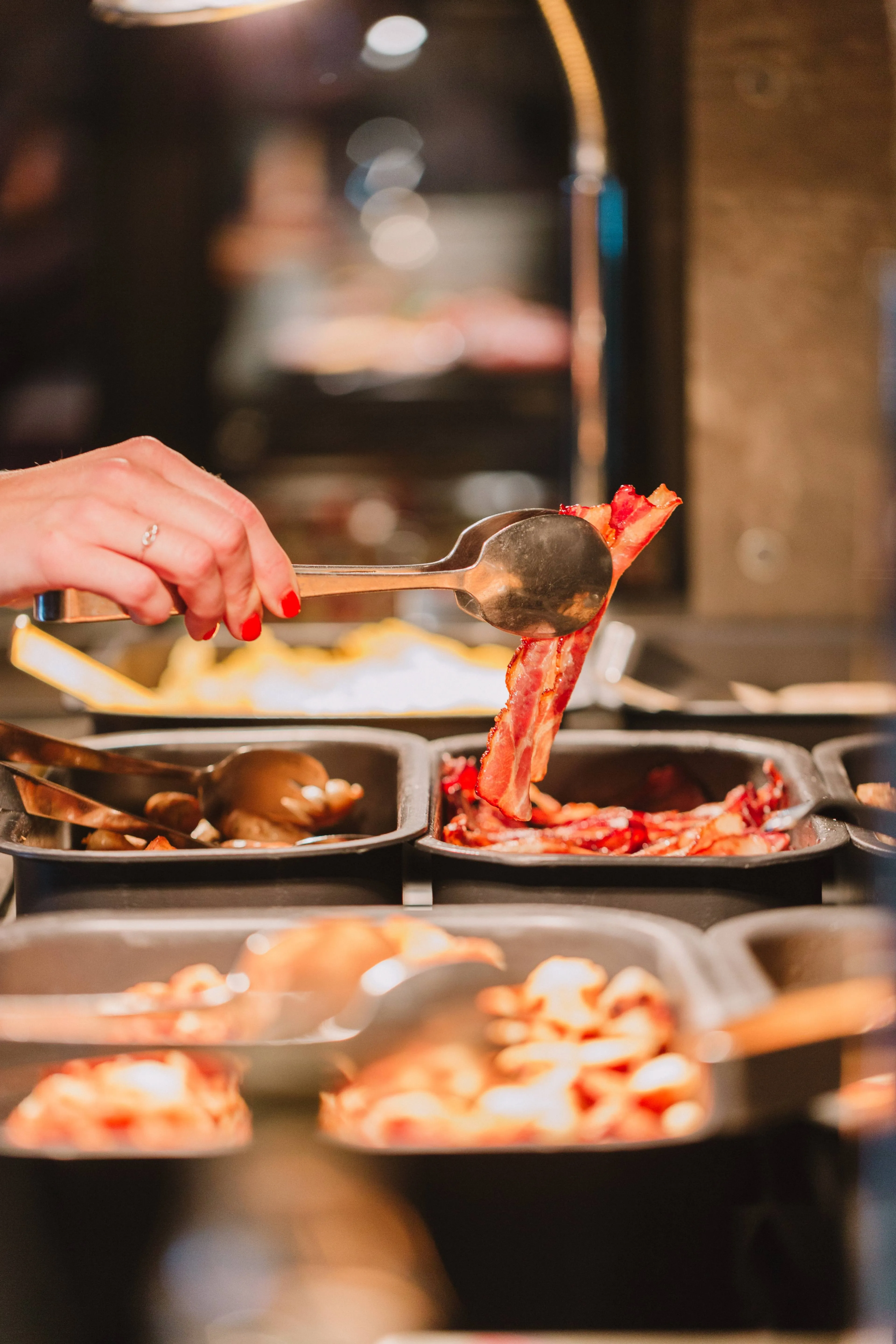 Eine Hand mit rotem Nagellack greift mit einer Zange nach frisch gebratenem Bacon – ein Blick auf das warme Frühstücksbuffet mit Eiern und Beilagen.
