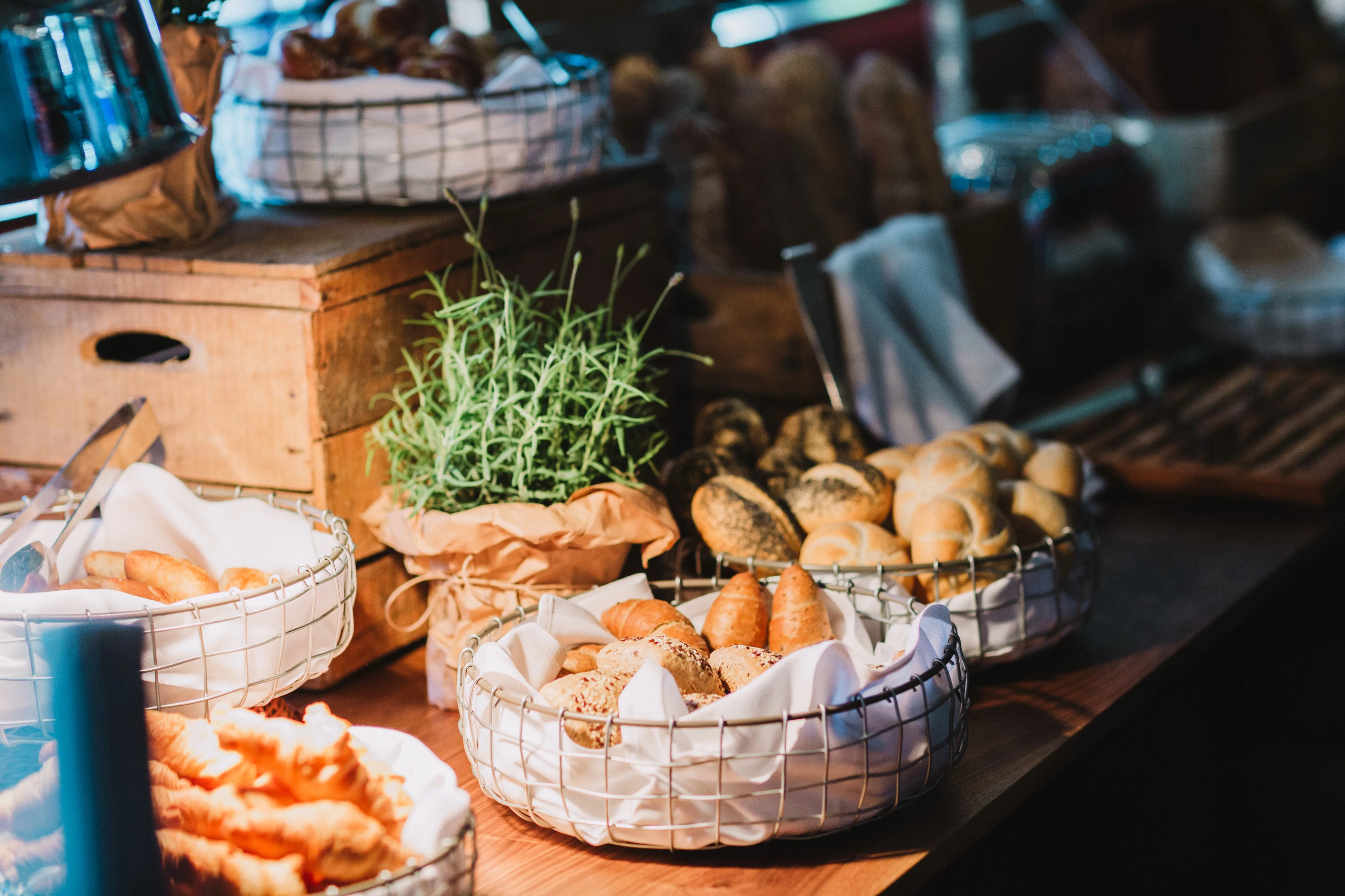 Verschiedene Körbe mit frischen Brötchen und Croissants auf einem Holztisch, umgeben von Kräutern und rustikaler Dekoration.