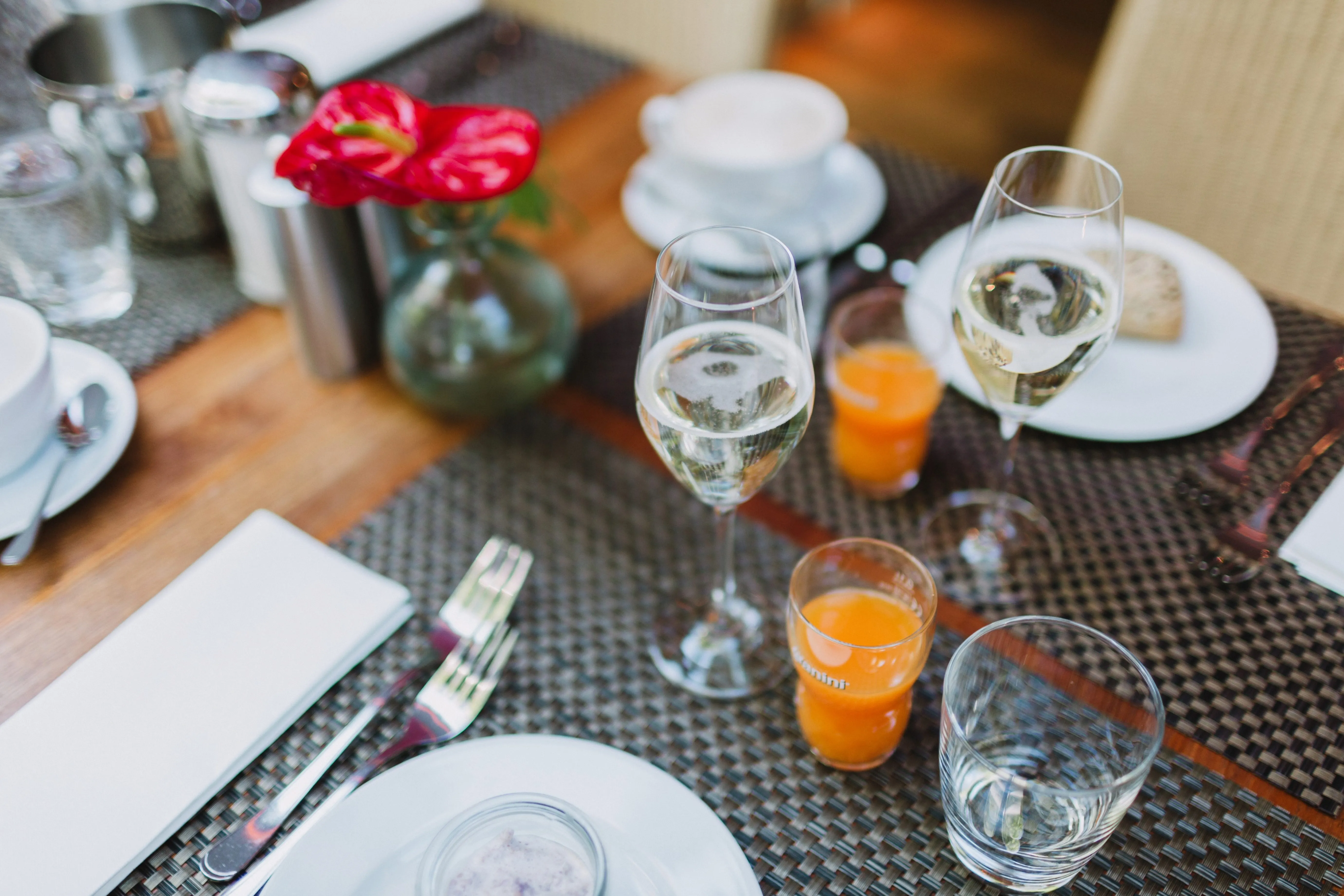 Elegant gedeckter Frühstückstisch mit Sektgläsern, Orangensaft, Wasser und einer roten Blume in einer Vase.