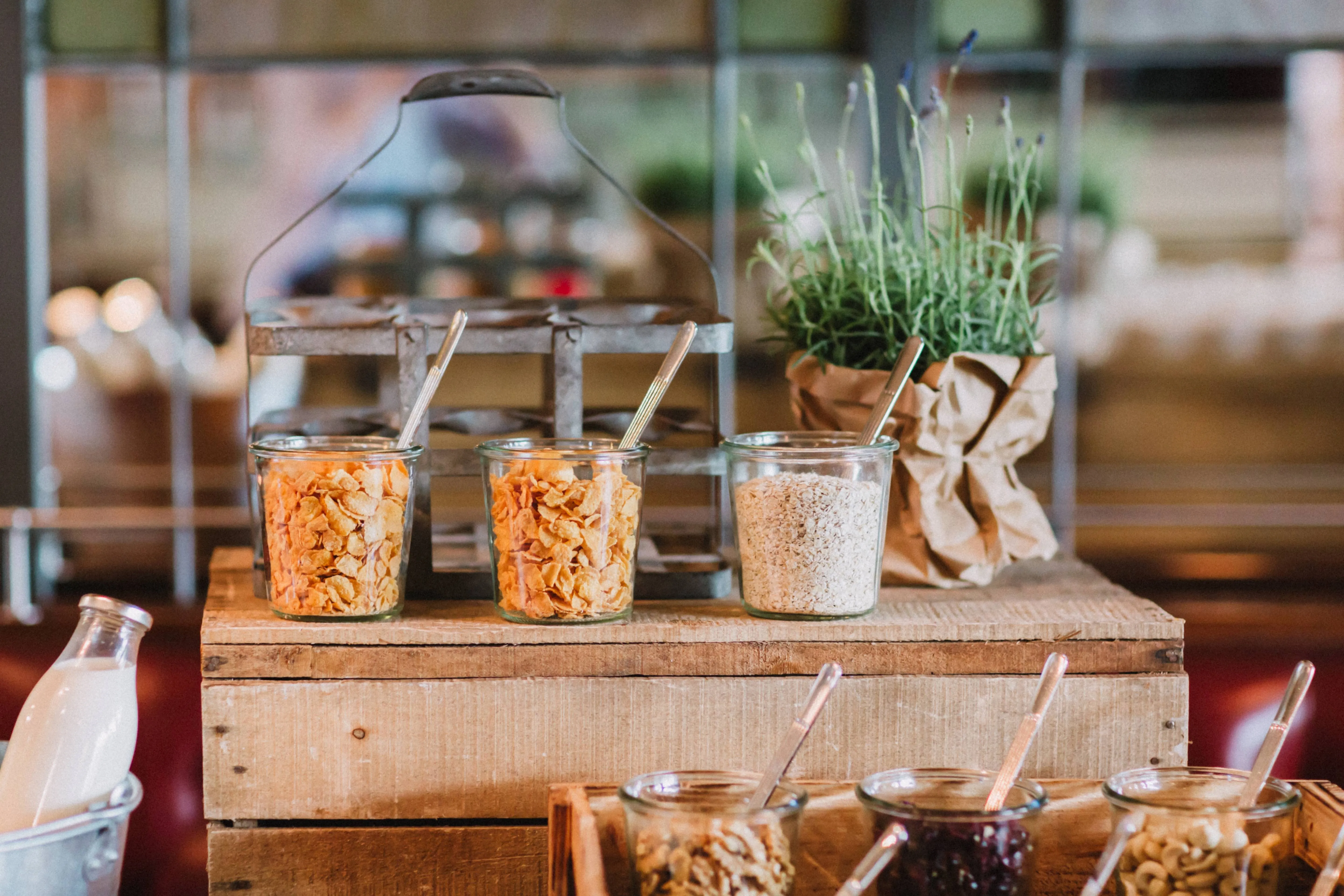 Buffet mit verschiedenen Müslisorten und Cornflakes in Glasbehältern, arrangiert auf Holzkisten – dazu eine Milchflasche und ein Kräutertopf als Deko.
