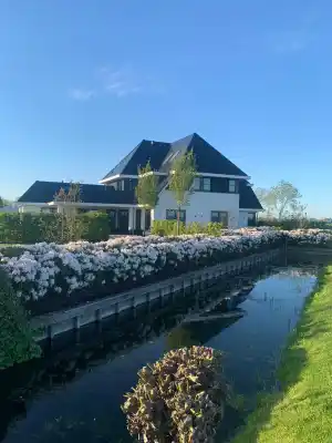 Modern white house with a dark roof next to a flowered hedge and a reflective water canal under a blue sky.