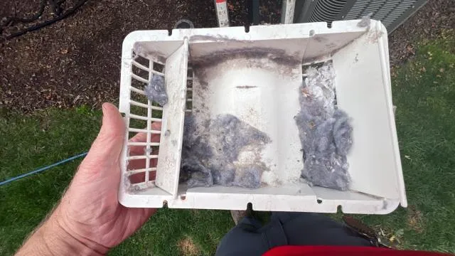 Dusty air conditioner filter held by hand outdoors near HVAC unit on grass.