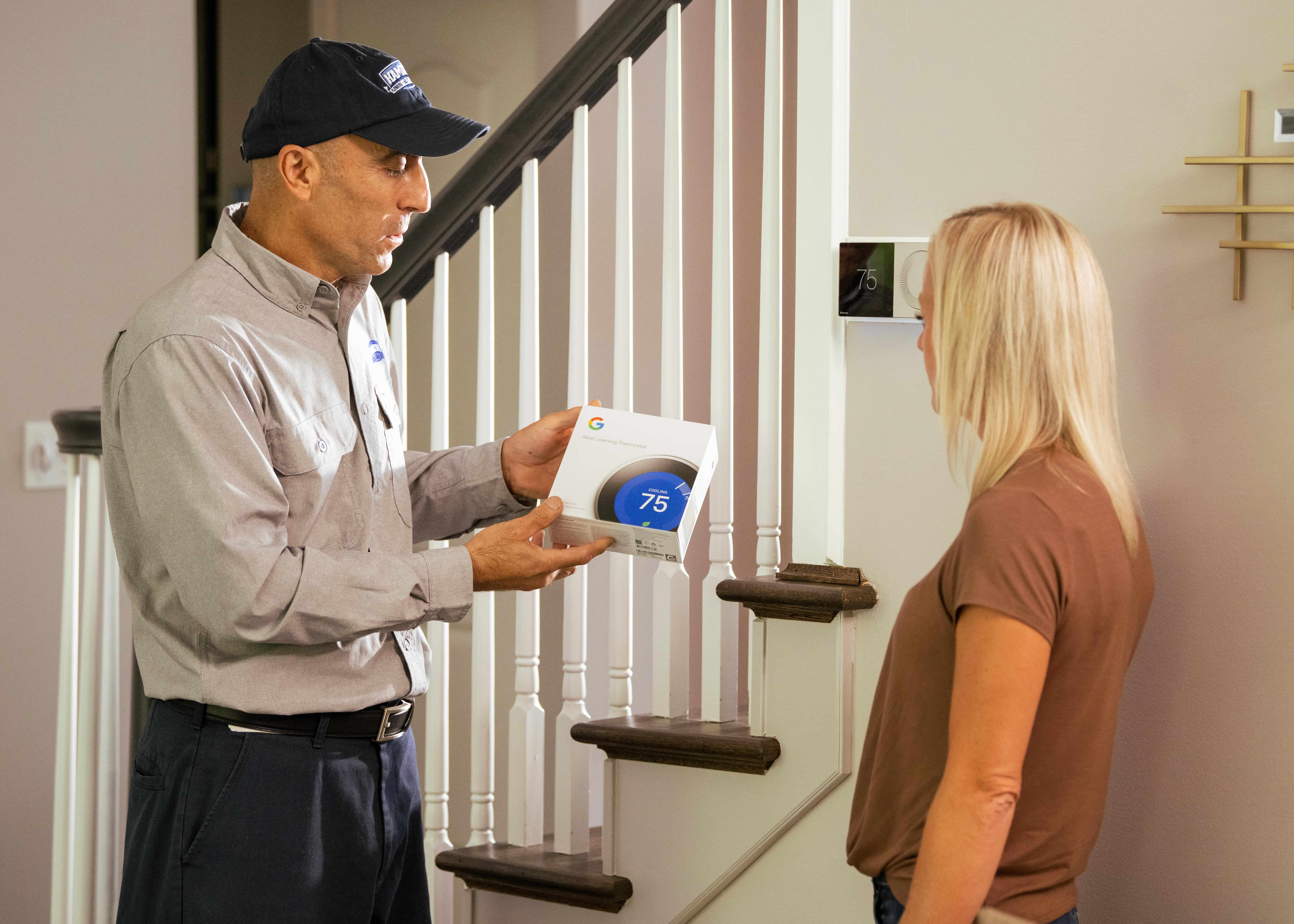 Technician shows client thermostat box, both standing next to a set of stairs and railing. The wall next to them has a digital thermostat installed.