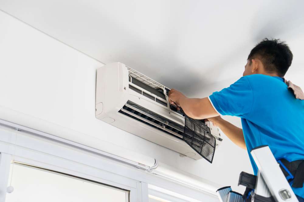 A technician stands on a ladder and works on a wall mounted ductless system, near the ceiling..