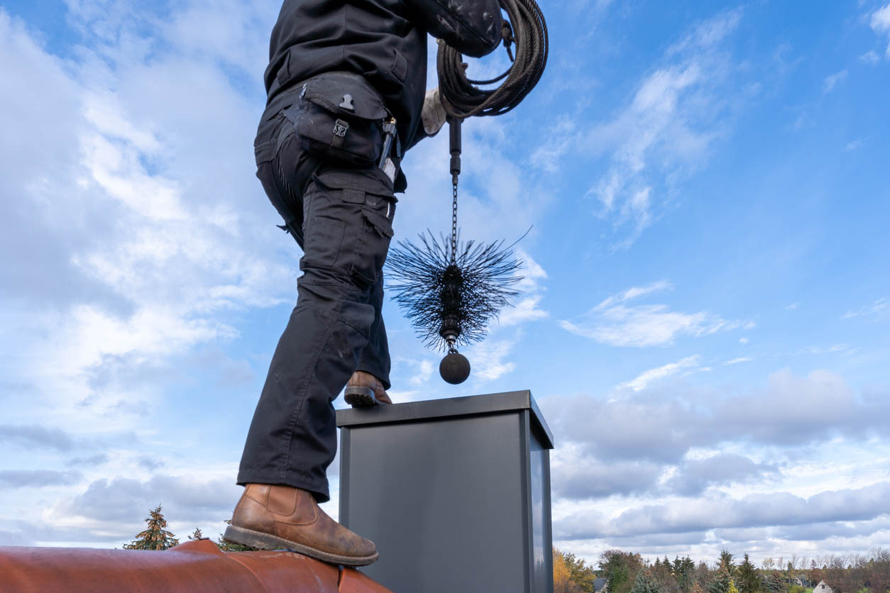 A technician stands outside on the roof by the chimney. He's holding a sweep brush above the chimney opening.