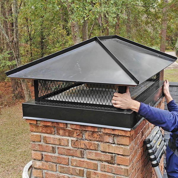 A technician standing on a ladder on a roof. His hands are installing a chimney cap.