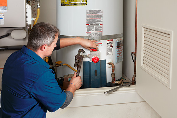 Technician in a blue uniform works on a water heater, holding a wrench to tighten a component.
