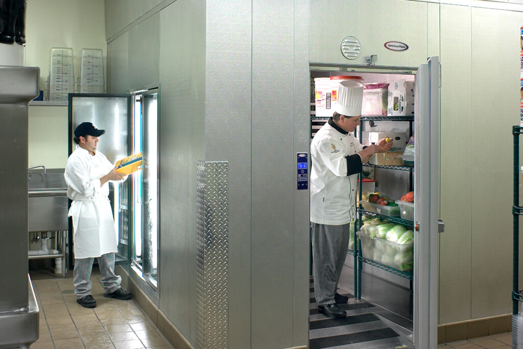 Image shows a chef checking product in a walking refrigerator, while another kitchen staff checks product from the refrigerator's reach in compartment.