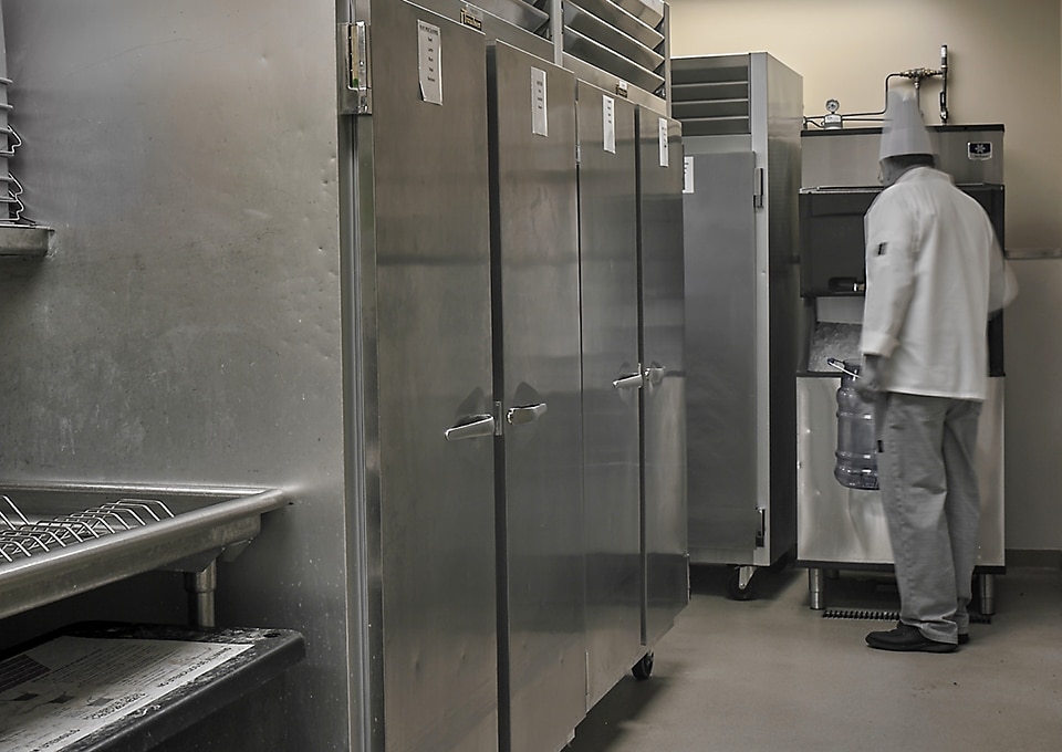 Image is of a set of commercial refrigerators in a restaurant. There's an employee scooping ice from an ice machine next to the refrigerators.