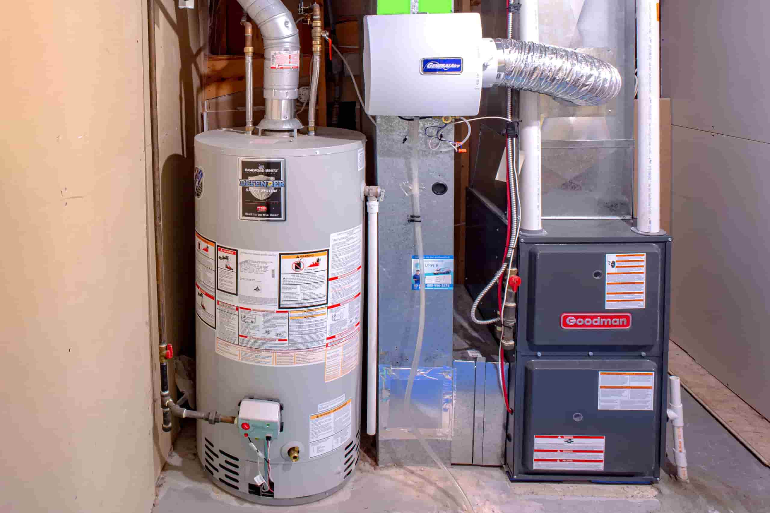 The image displays a home heating and water heating system, specifically featuring a Goodman furnace and a water heater.