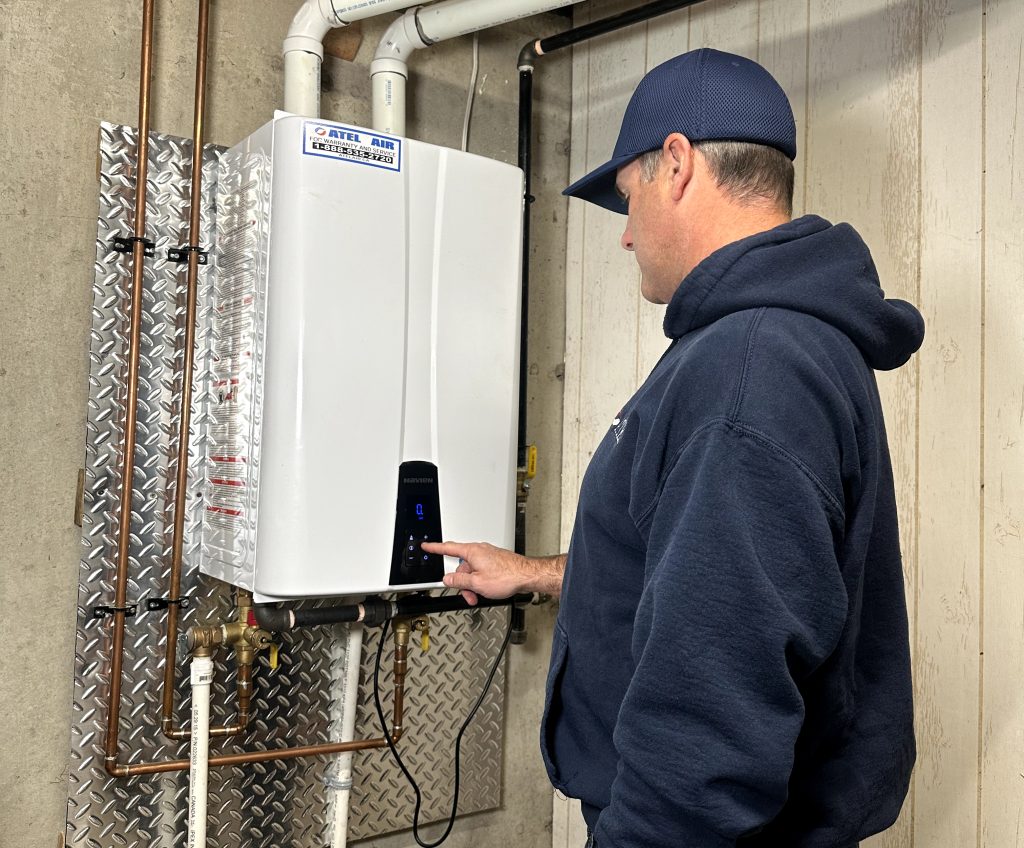 Un hombre con una capucha negra y una gorra azul está revisando un calentador de agua sin tanque.