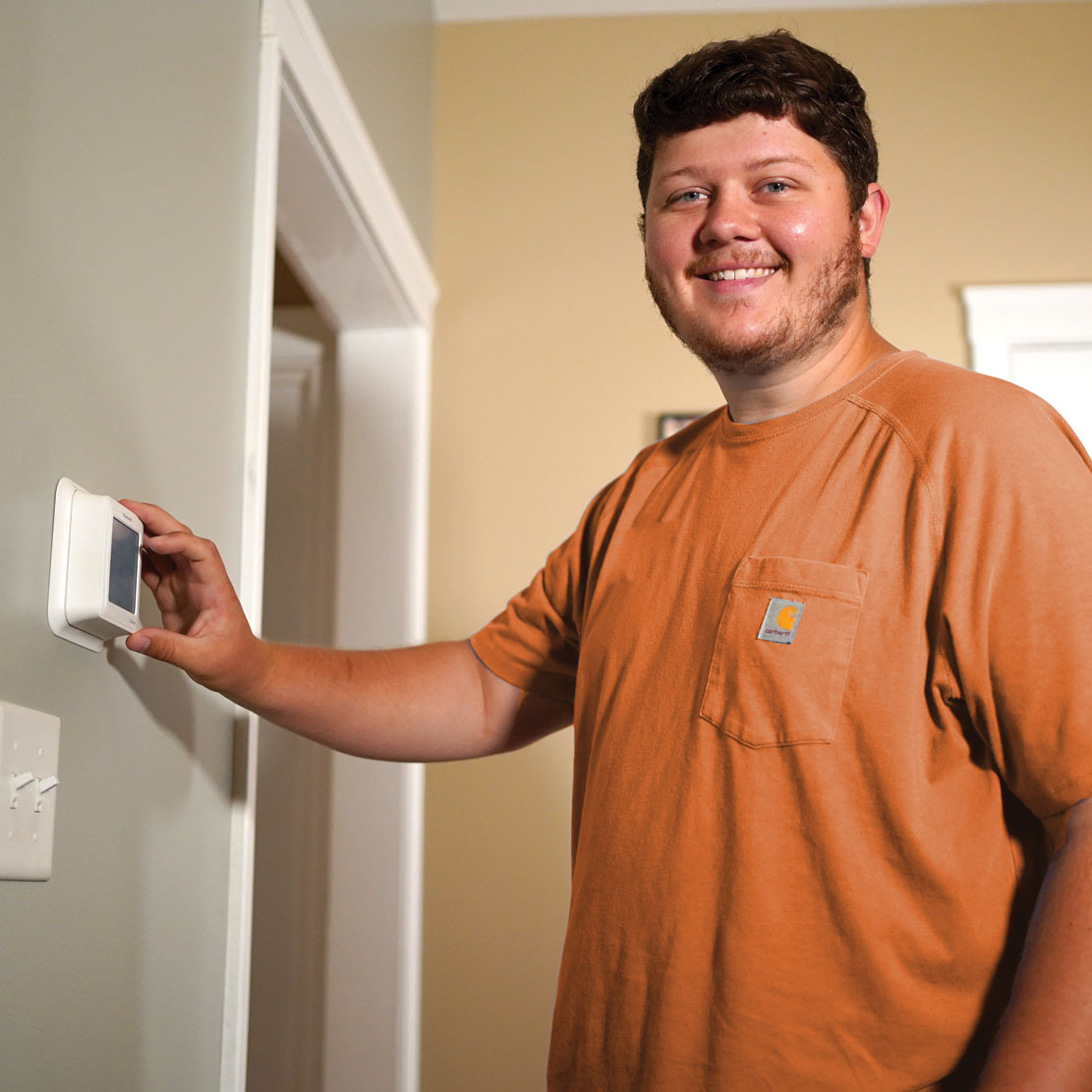 Hombre con una camisa naranja revisa un termostato digital en una pared, junto a una puerta.